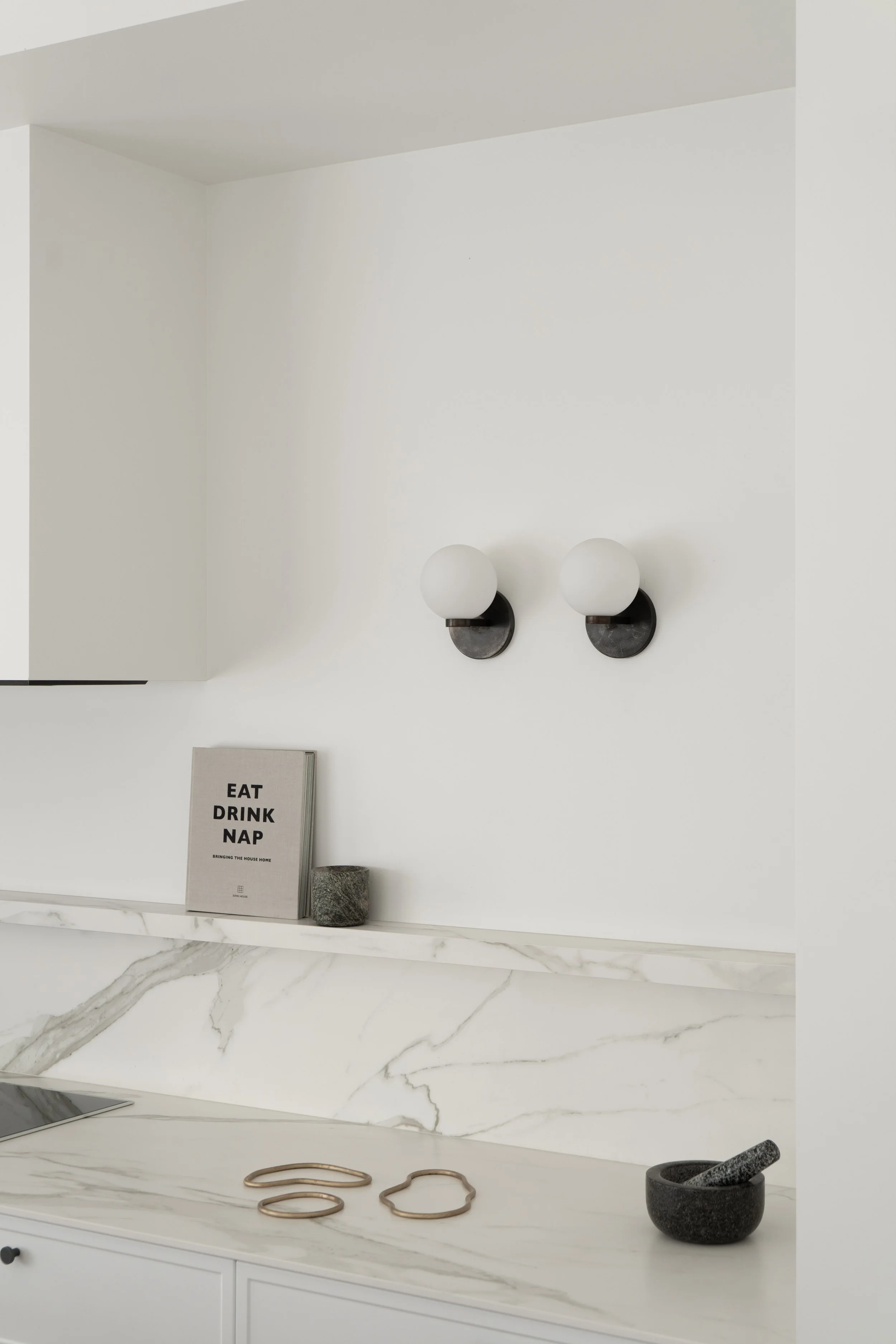 A white kitchen shelf with a marble countertop holding jewelry, a black mortar and pestle, and a sign that says "Eat Drink Nap". Above are two wall-mounted black and white spherical light fixtures.
