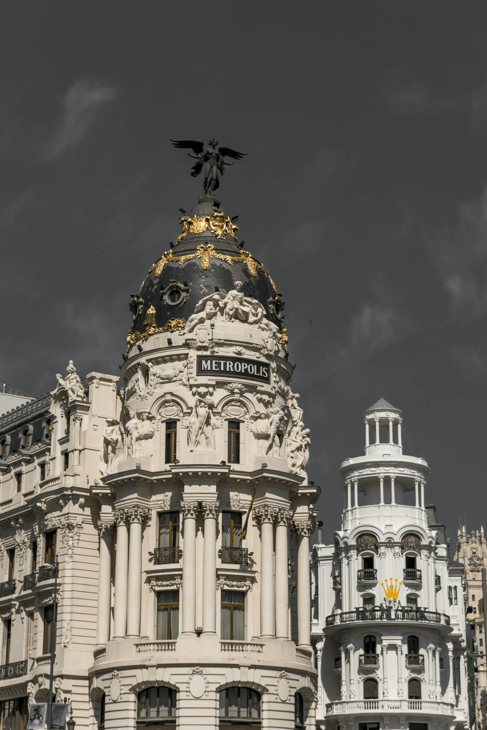 The Metropolis building in Madrid, Spain, featuring a white ornate facade with sculptures and a clock, topped with a black dome with gold accents and a winged statue.