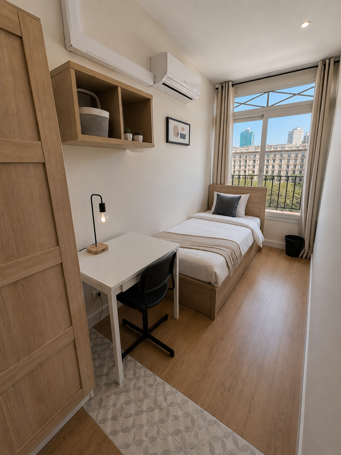 A small bedroom with a single bed, a desk with a black chair, and a window with city buildings outside. The room has wooden furniture and light-colored walls.