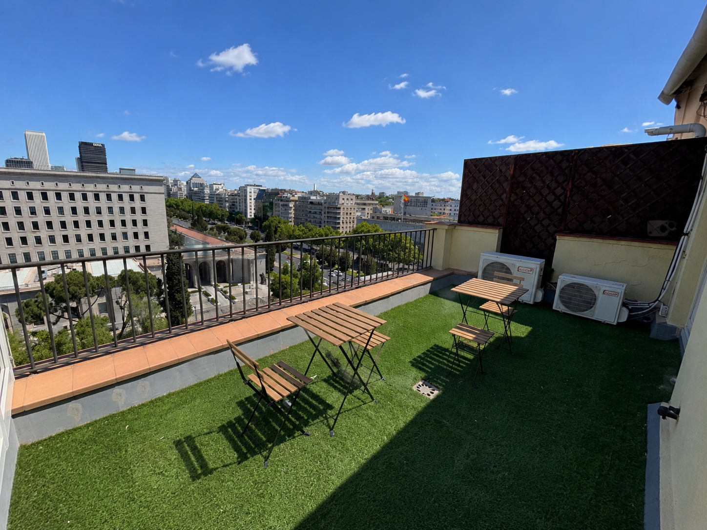 Rooftop terrace with artificial grass, small wooden tables, and chairs, overlooking city buildings and a clear blue sky with scattered clouds.