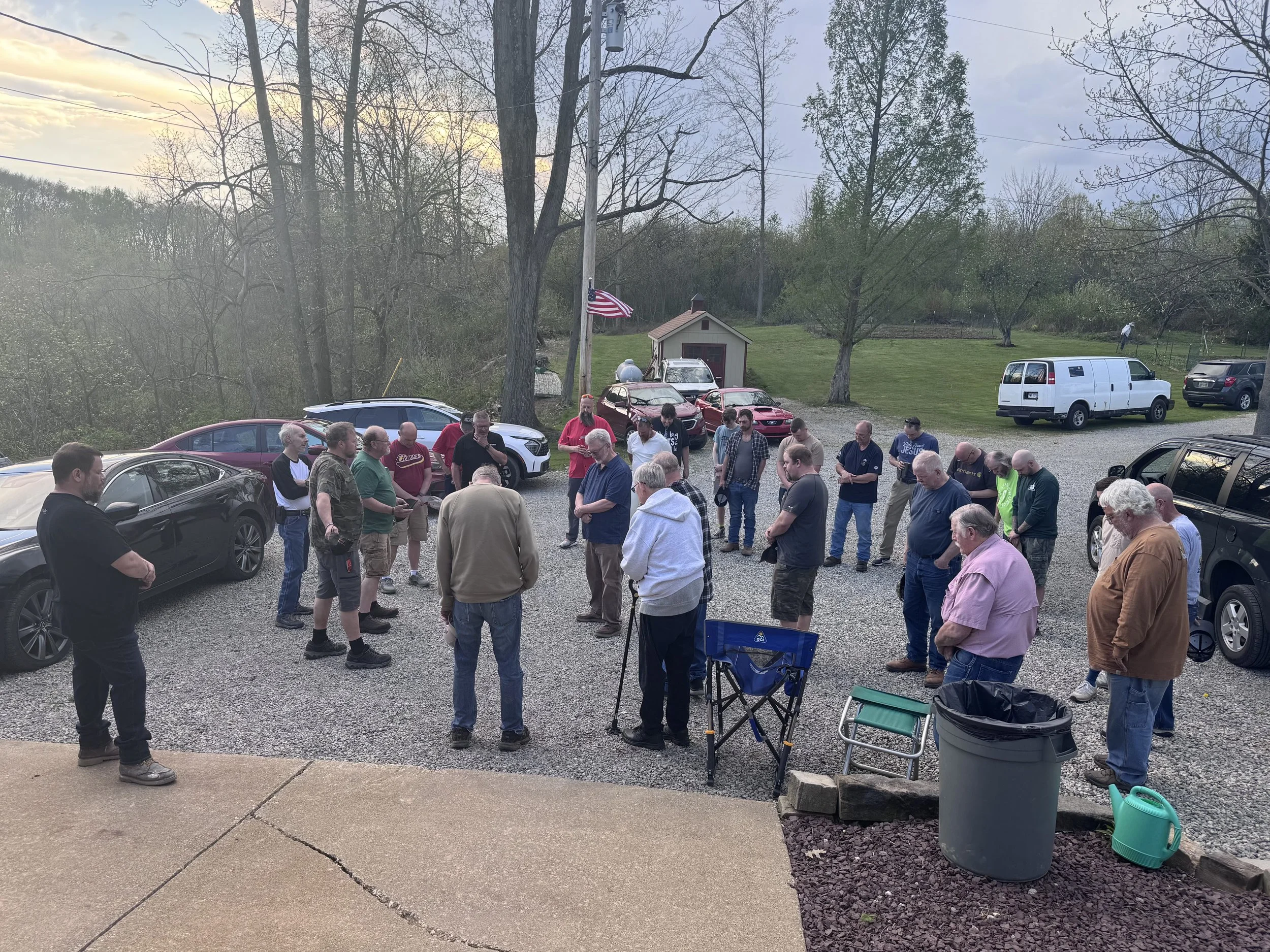 A group of people gathering outdoors in a driveway or yard with several parked cars and trees in the background, possibly during a community or social event.