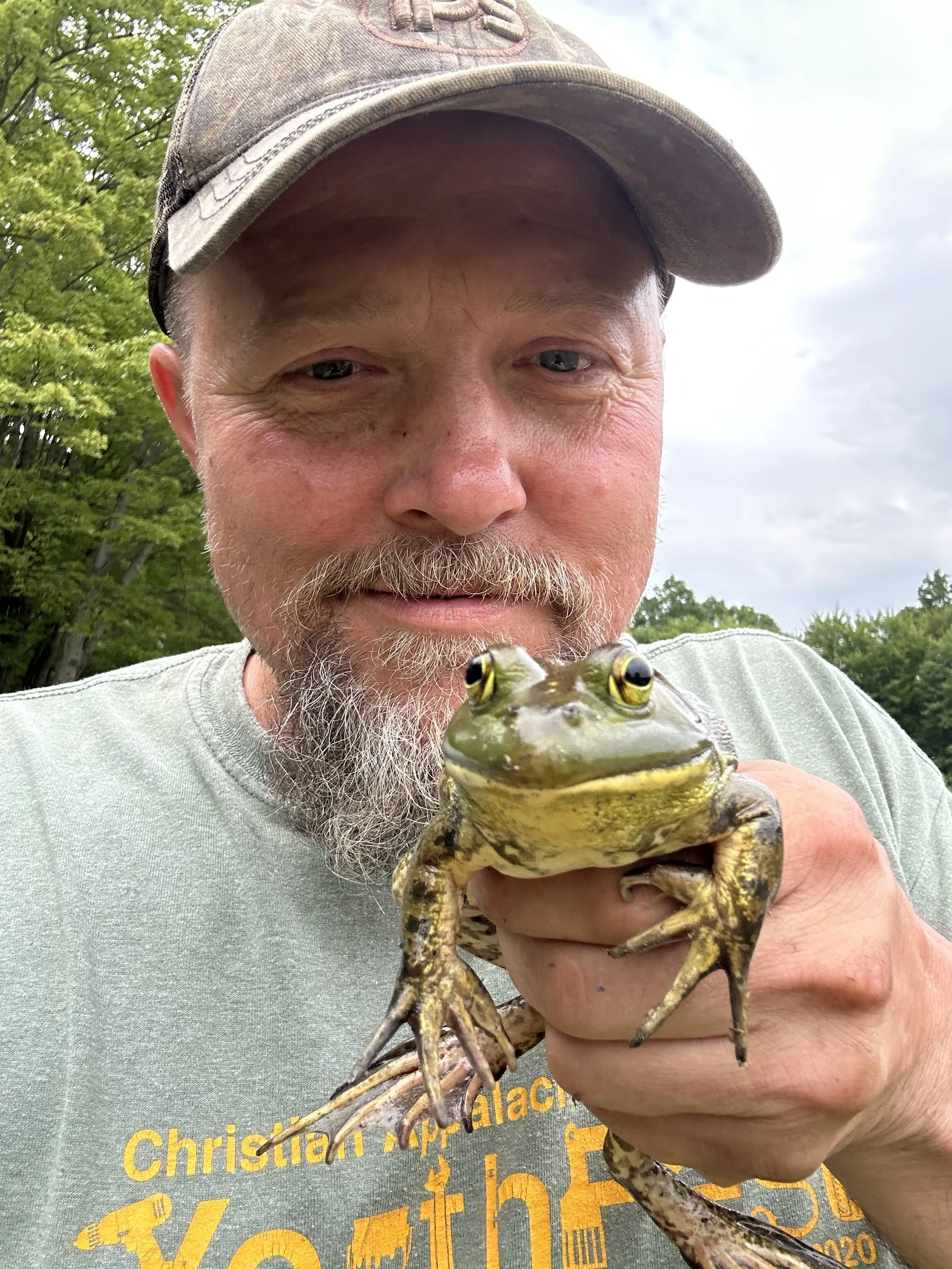 A man outdoors holding a frog close to the camera with trees and sky in the background.