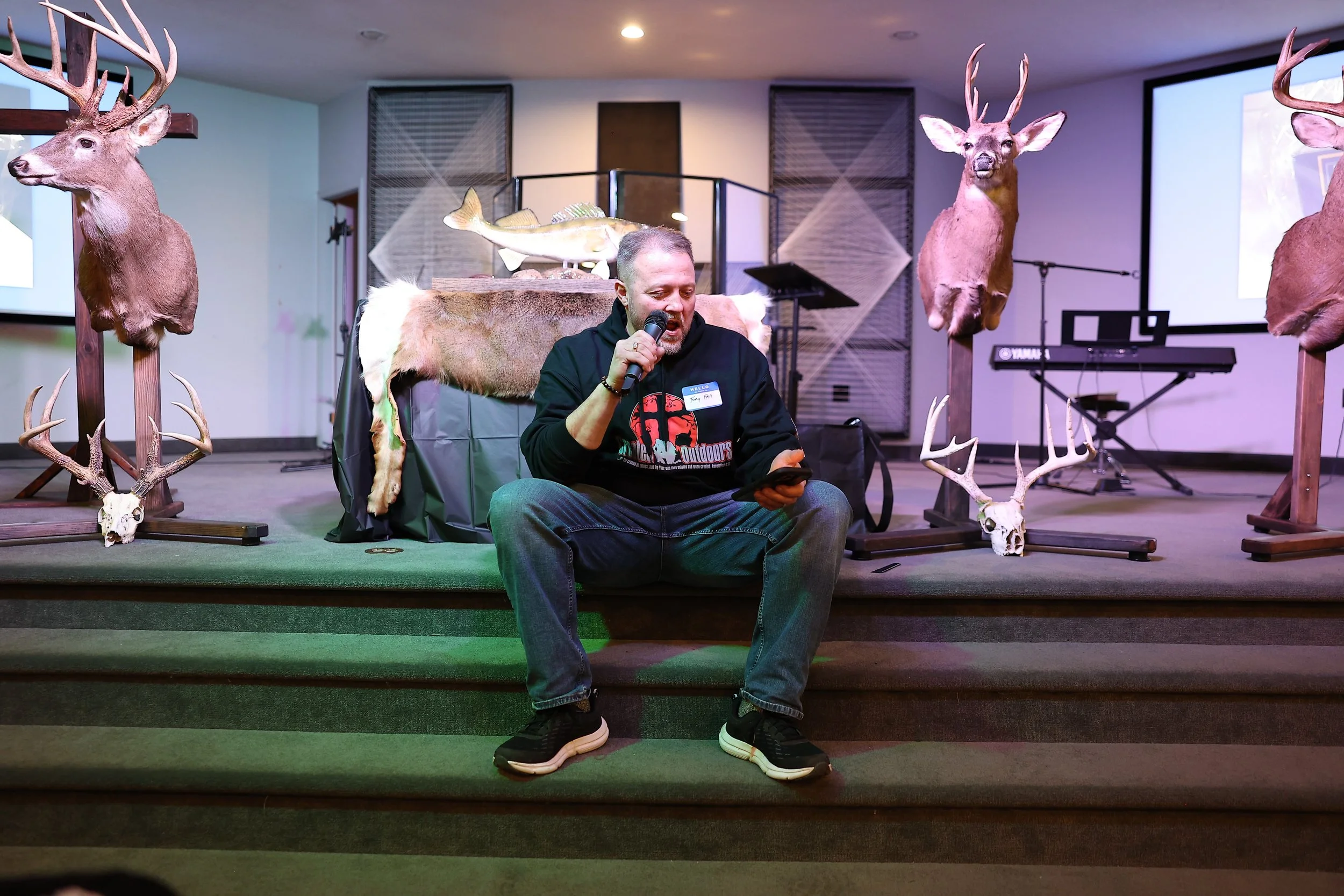 A man sitting on a stage with mounted animal heads and fur displays, speaking into a microphone.