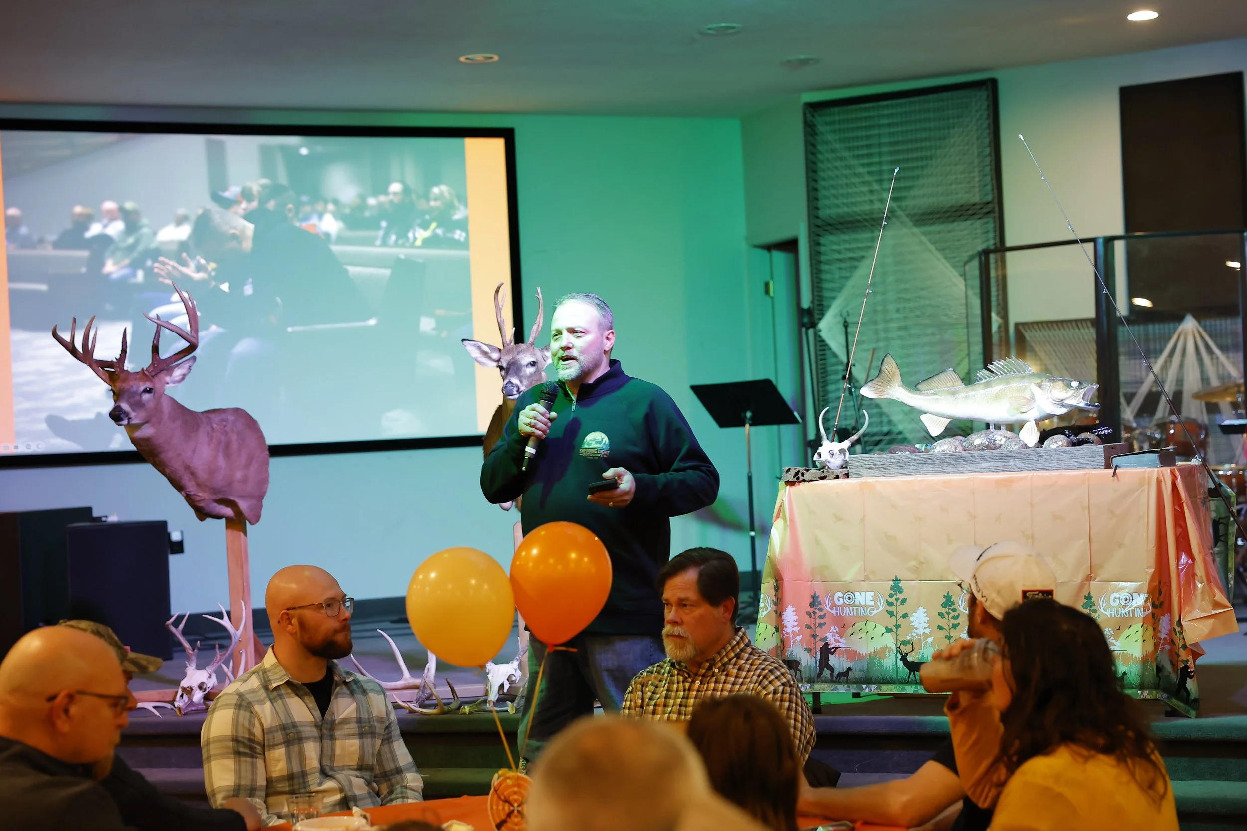 A man speaking into a microphone at a hunting-themed event, with deer and fish decorations, and a large screen displaying an audience in the background.