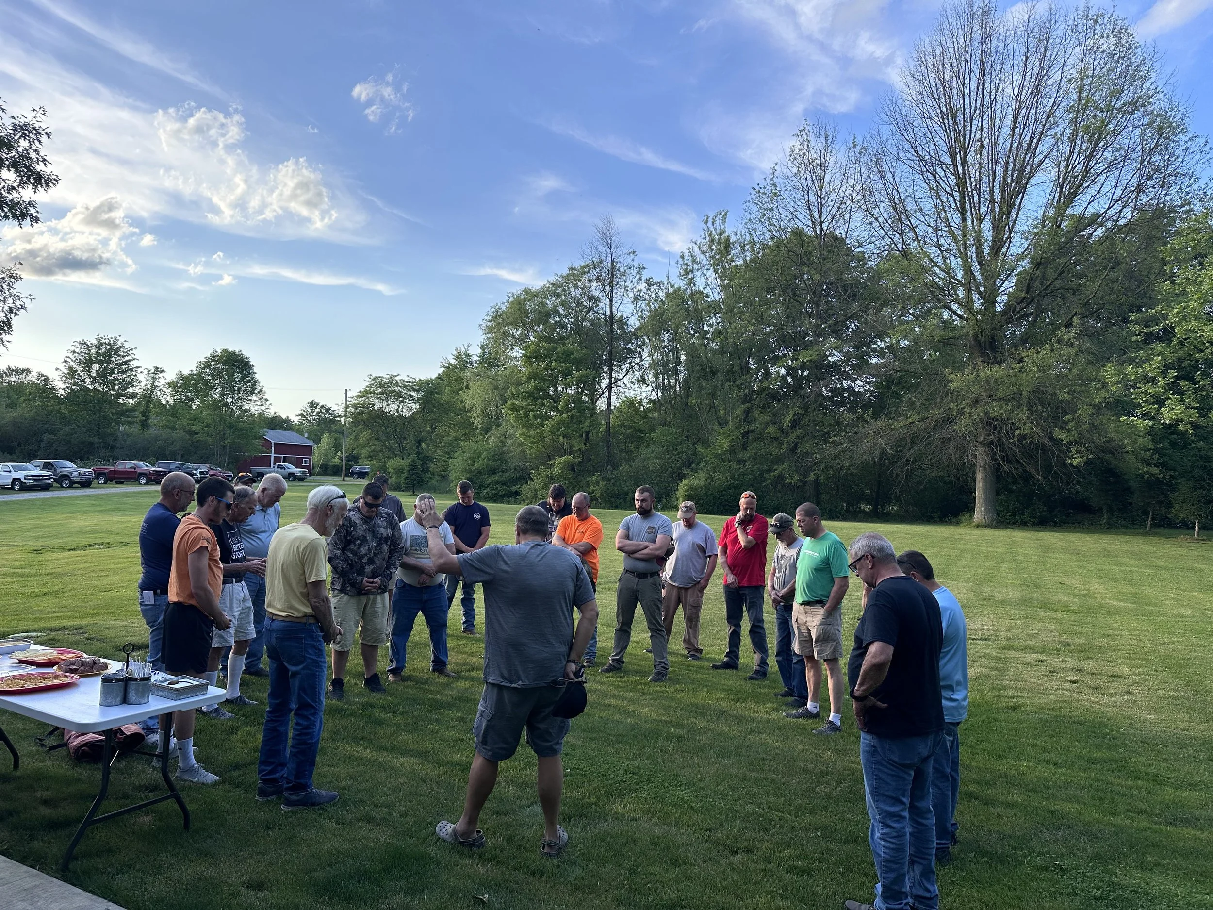 A group of people standing outdoors in a grassy area during the day, possibly participating in a prayer or moment of silence, with a table of food nearby, trees in the background, and a partly cloudy sky.