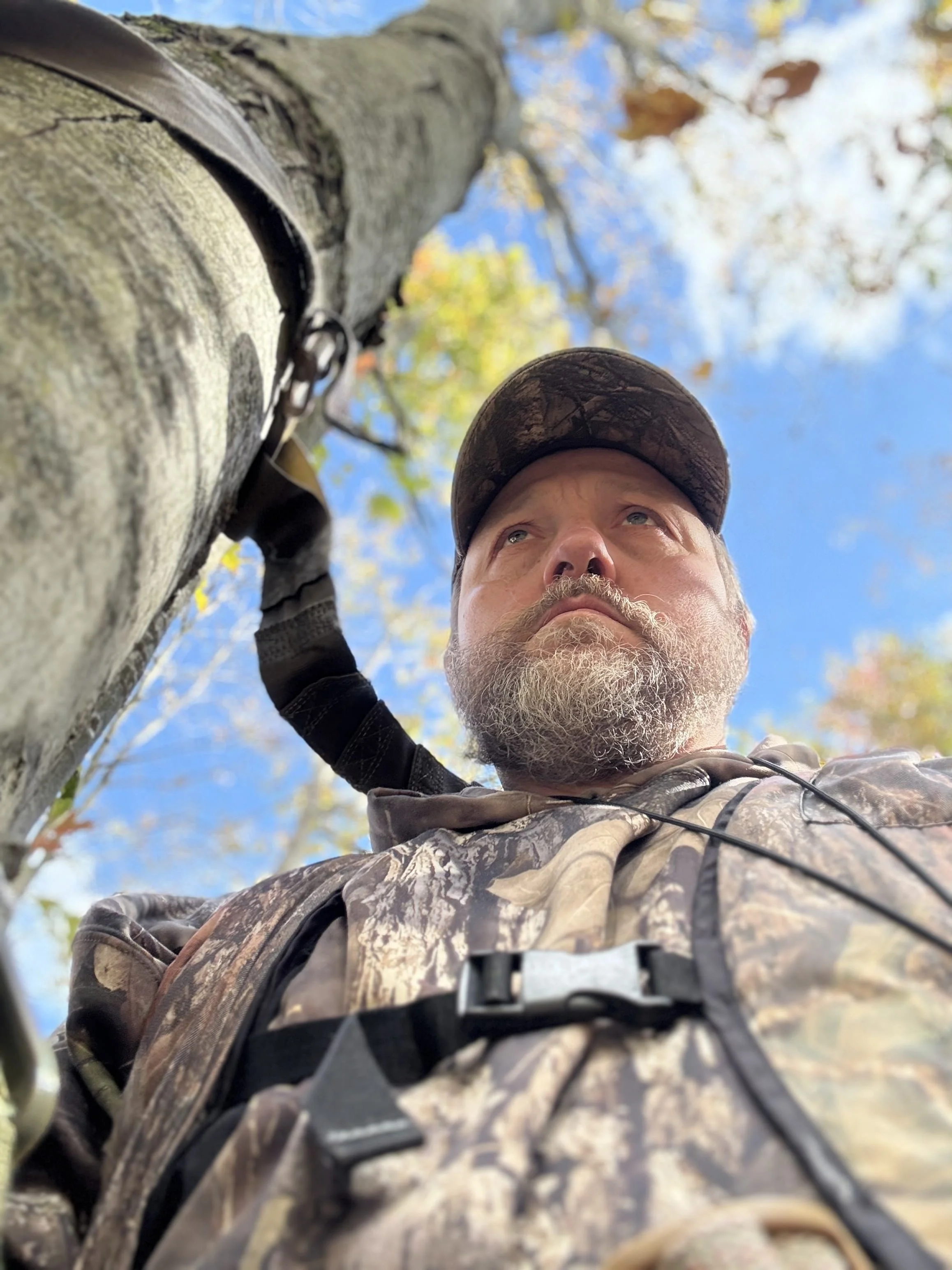 Close-up of a man wearing camouflage clothing and a cap, looking up at a tree with fall foliage.