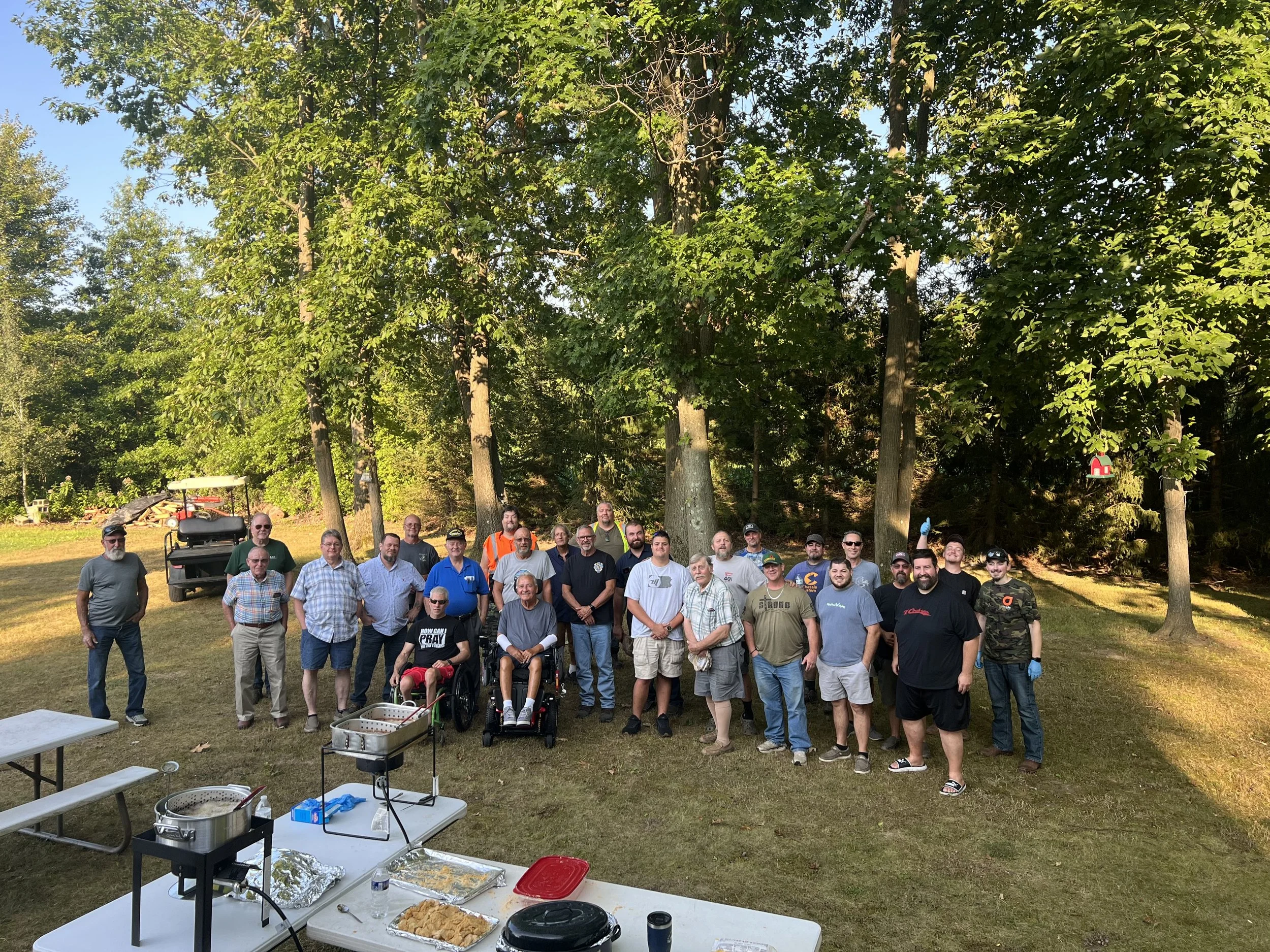 A group of people gathered outdoors in a park with tall trees in the background. There is a table with food and supplies in the foreground. The group includes men and women of various ages, some wearing casual clothing and others in uniforms or work 