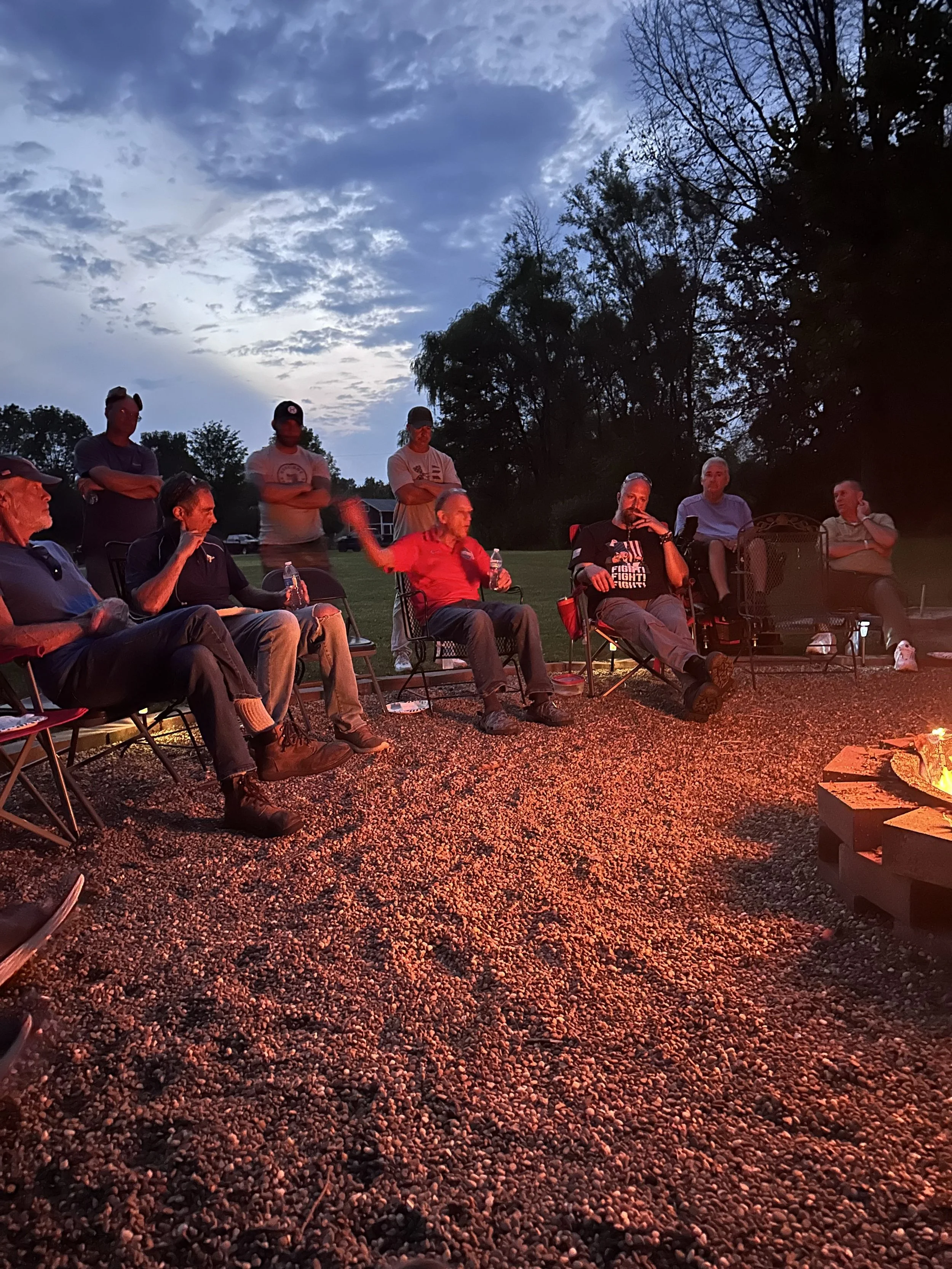 A group of people sitting in chairs around a campfire outdoors during twilight or early evening, with trees and a cloudy sky in the background.