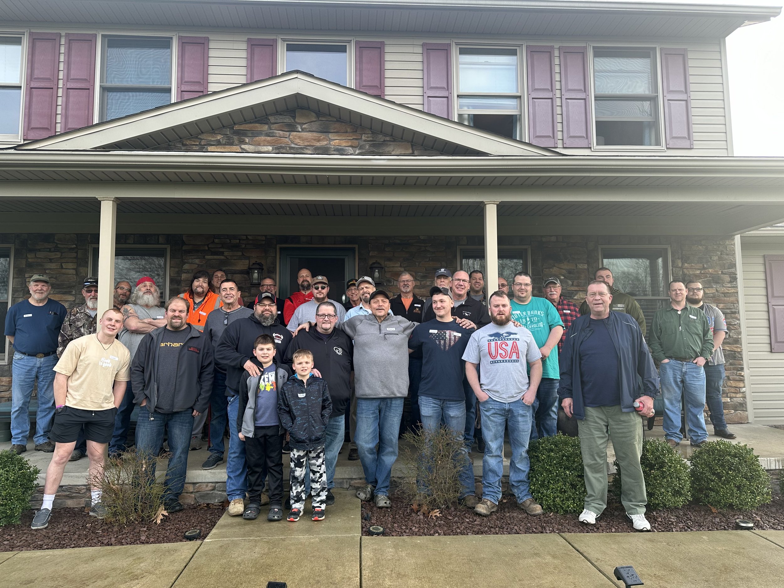 A large group of people, including men, women, and children, are gathered on the front porch of a house with gray siding and purple shutters. The house has a stone accent on the gable. The group is posing for a photo and appears to be smiling.