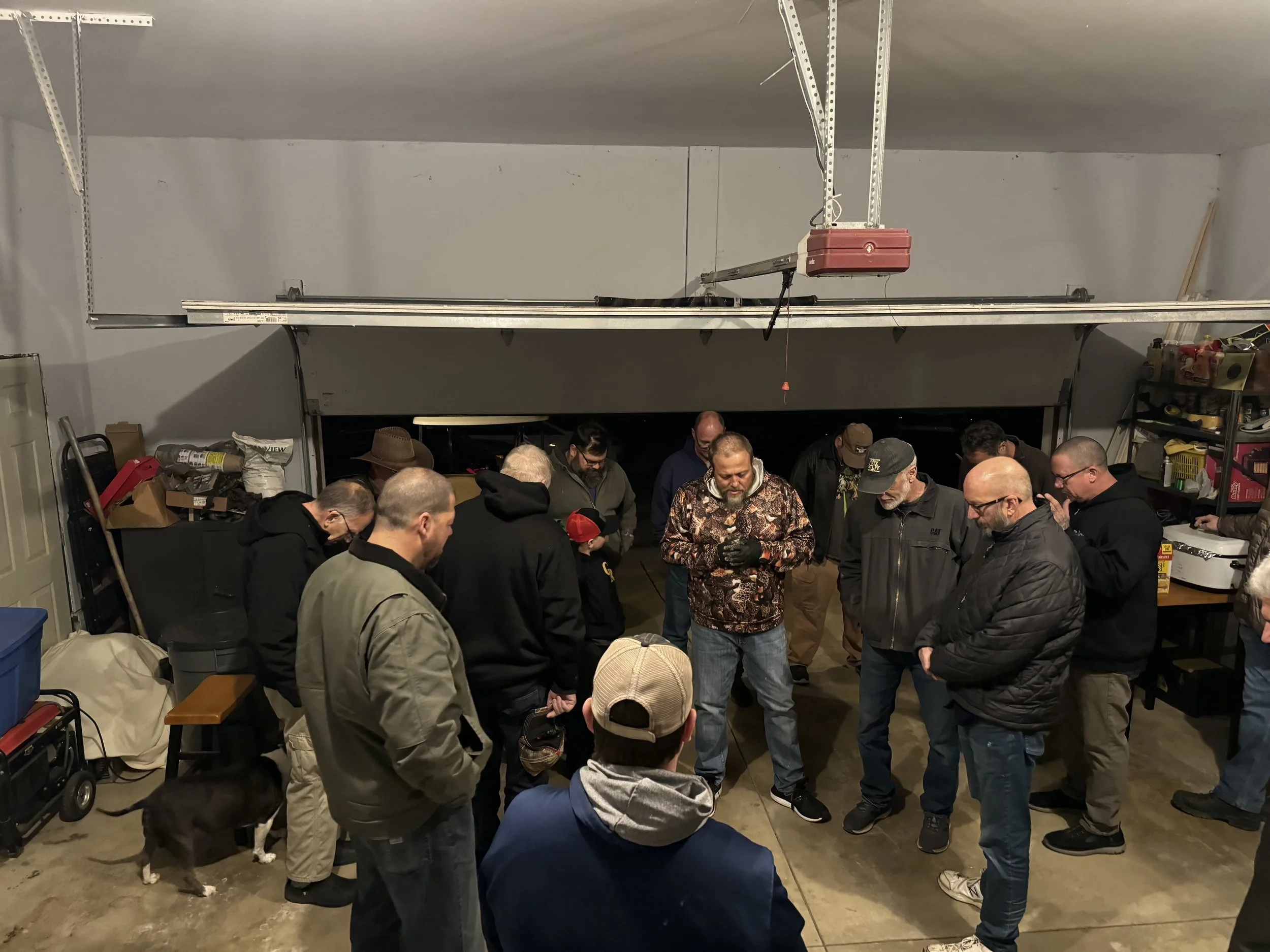 A group of people gathered in a garage, some standing and some kneeling, with various tools and boxes around. The garage has a high ceiling with visible pipes and a garage door on the left. The people appear to be engaged in a group activity or discu