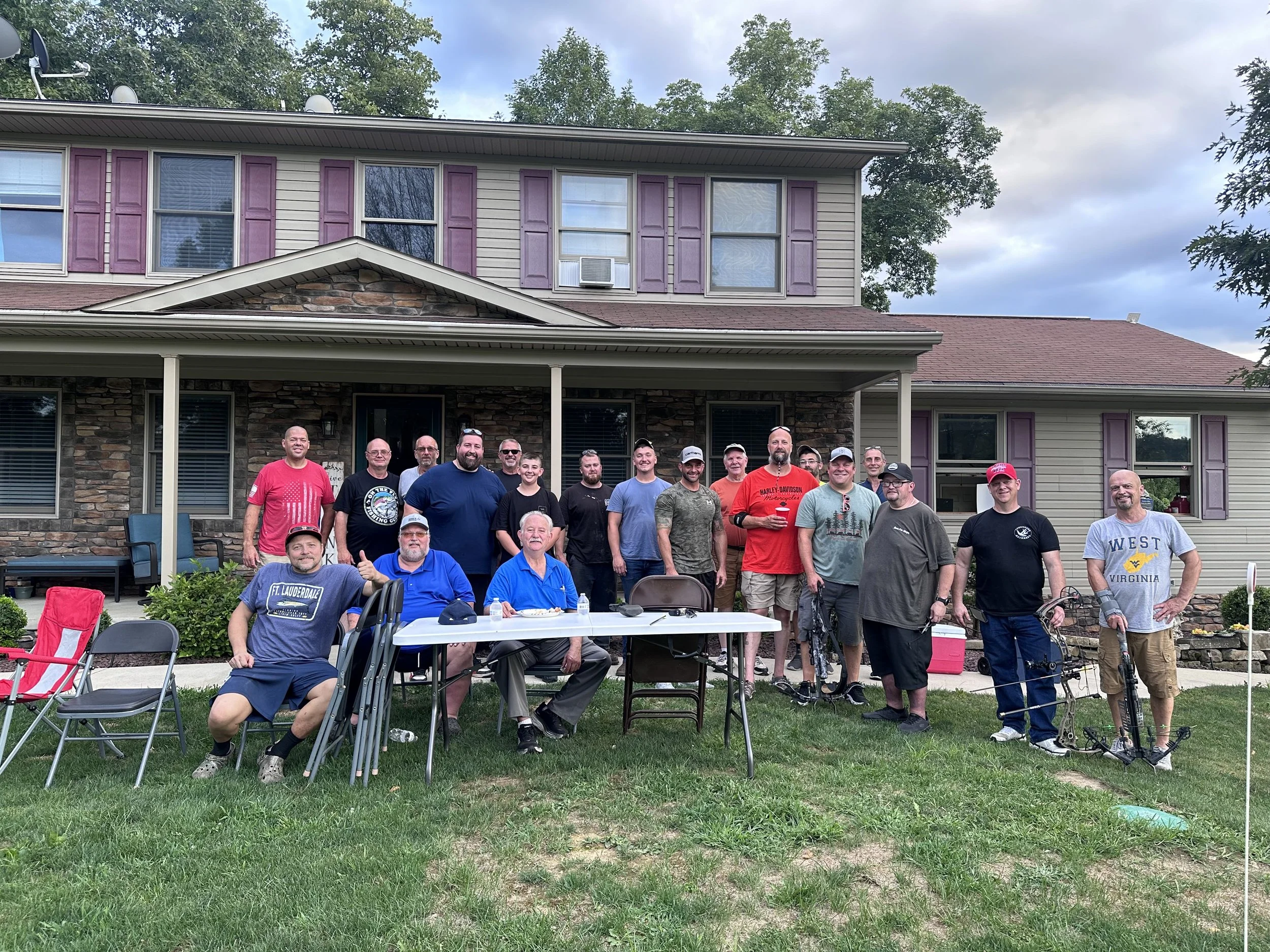 Group of people gathered outdoors in front of a house, some seated at tables and others standing, with trees and cloudy sky in the background.