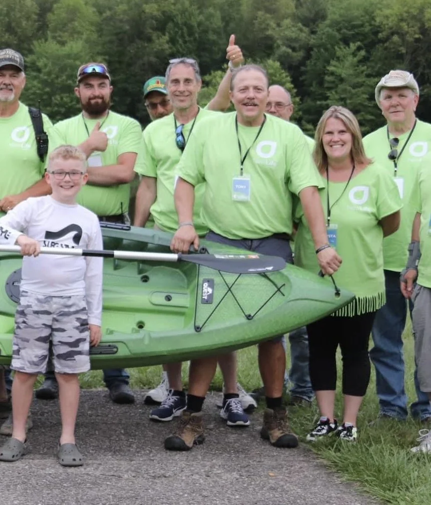 Group of people, some wearing matching neon green T-shirts, standing outdoors with a green kayak and paddles. They are smiling, with one person giving a thumbs up or raising a hand, in a natural wooded area.