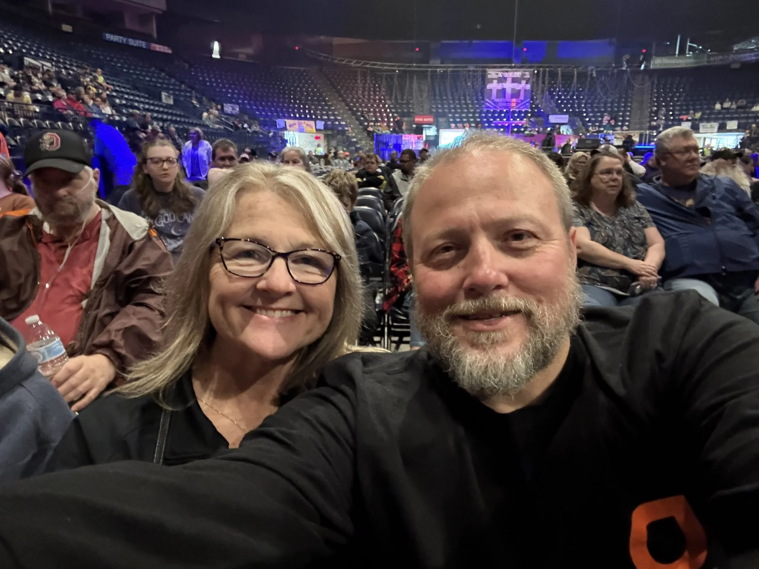 A group of people seated in an indoor arena or concert venue, with two people in the foreground taking a selfie, showing their smiling faces. The background displays a stage with lights and screens, and a mostly empty seating area.