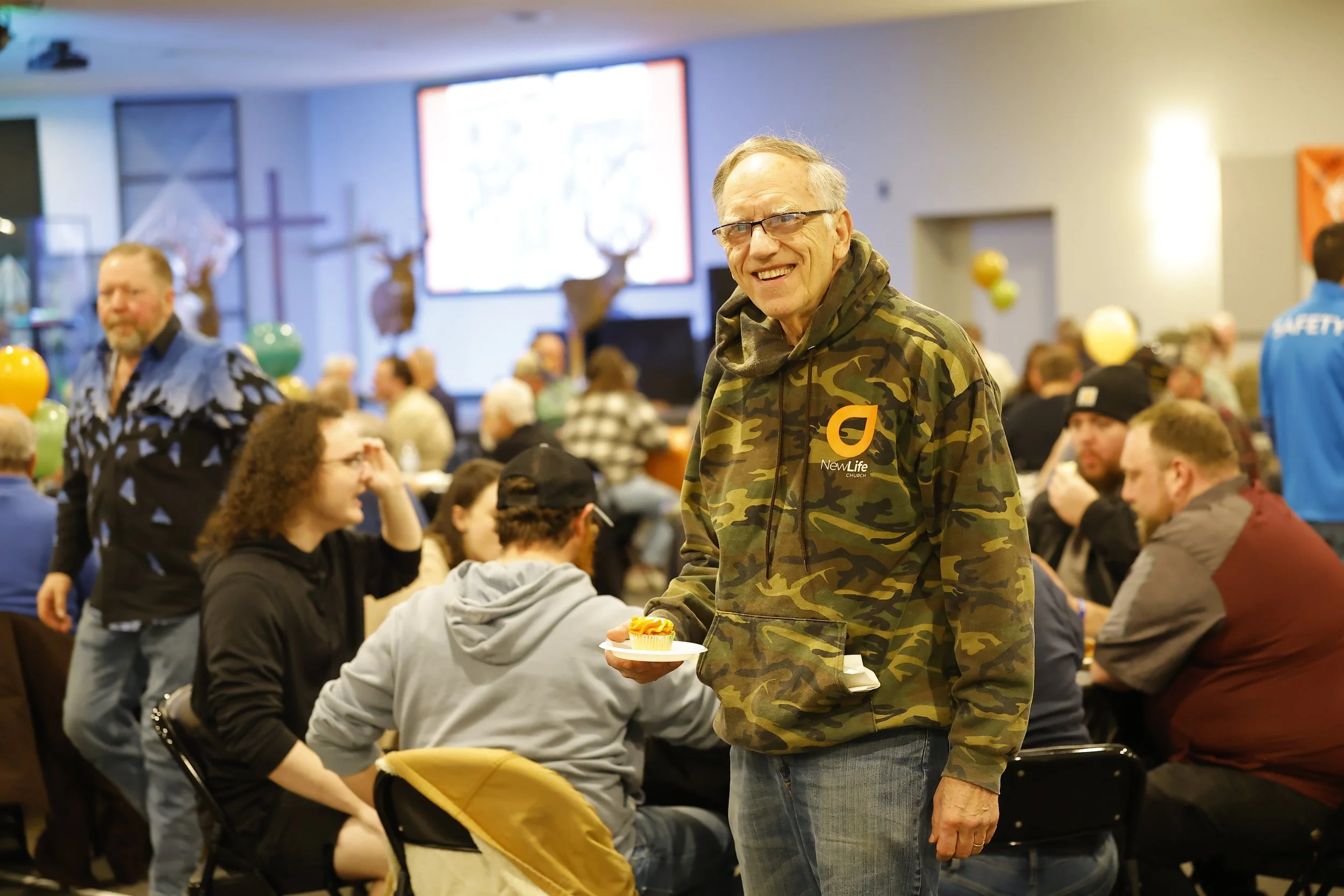 An elderly man in glasses and a camo jacket with a 'New Life Church' logo is smiling while holding a cupcake on a plate. He is in a large room filled with people, some seated at tables and others standing, engaging in a community event. The room is decorated with balloons and mounted animal heads, and a large screen is visible in the background.