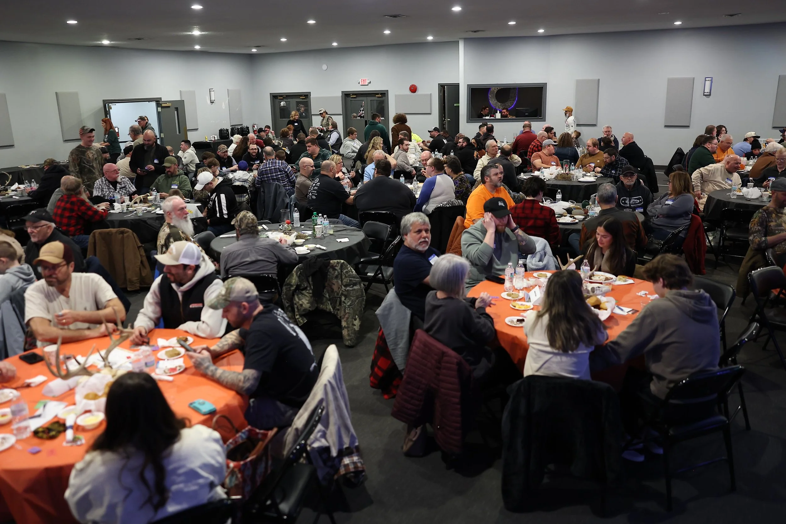 A large indoor gathering with many people seated at round tables with orange and black tablecloths, eating and socializing. Some are wearing casual clothes, hats, and jackets. There are food and drinks on the tables, and more people are walking around or standing in the background. The room has gray walls, a few doors, and mounted screens or panels.