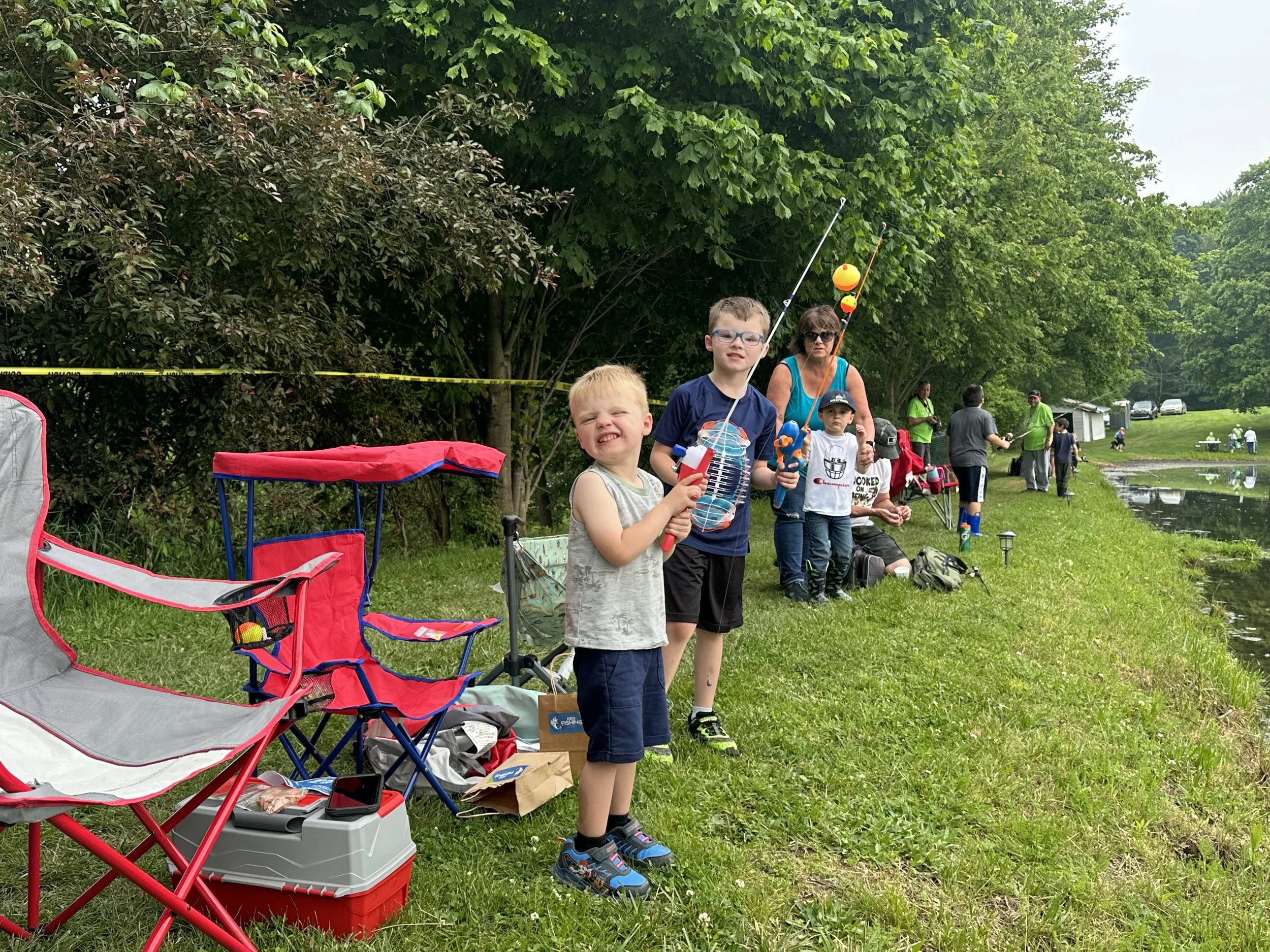 A group of children and adults fishing by a lake on a grassy bank, with trees and cars in the background, and fishing equipment and chairs in the foreground.