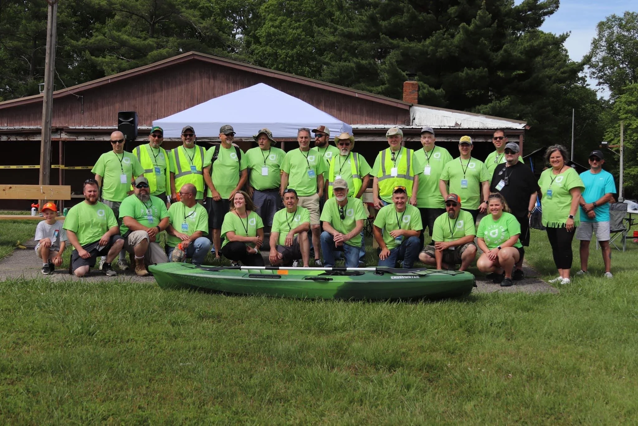 A group of people wearing matching bright green shirts gathered outdoors in front of a rustic building with a white tent set up behind them. They are smiling and posing for a group photo, with a kayak in the foreground.