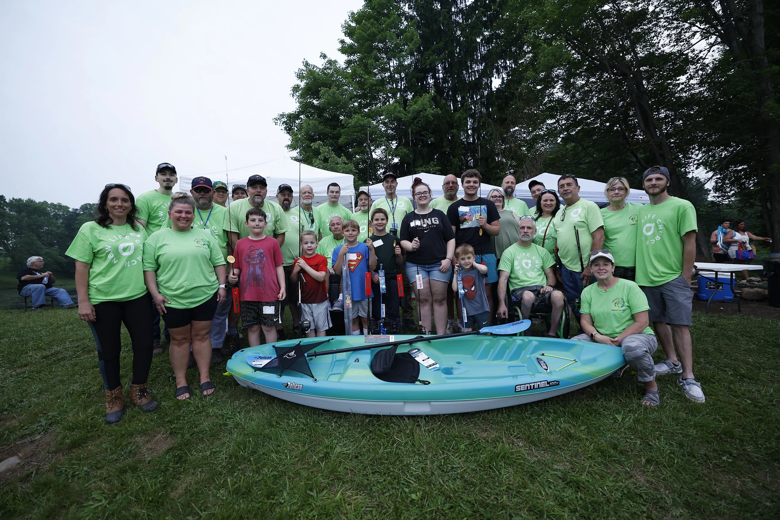 Group of people, including children and adults, gathered outdoors on grass with trees and tents in the background, posing for a photo. Some are wearing matching bright green T-shirts, and there are kayaks and a paddleboard in front.
