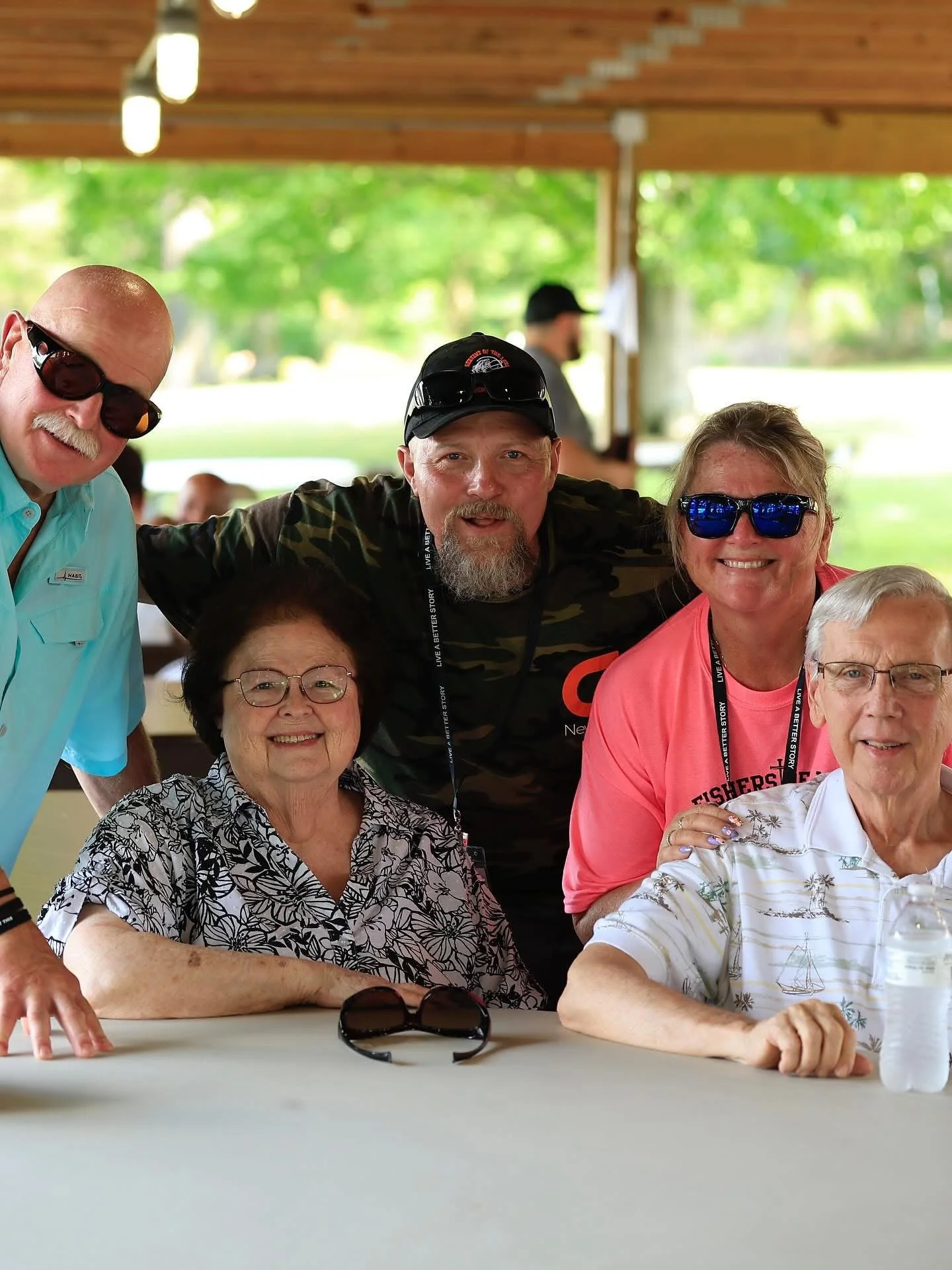 Group of five smiling older adults sitting at a table outdoors with greenery in the background, some wearing sunglasses and casual clothing.
