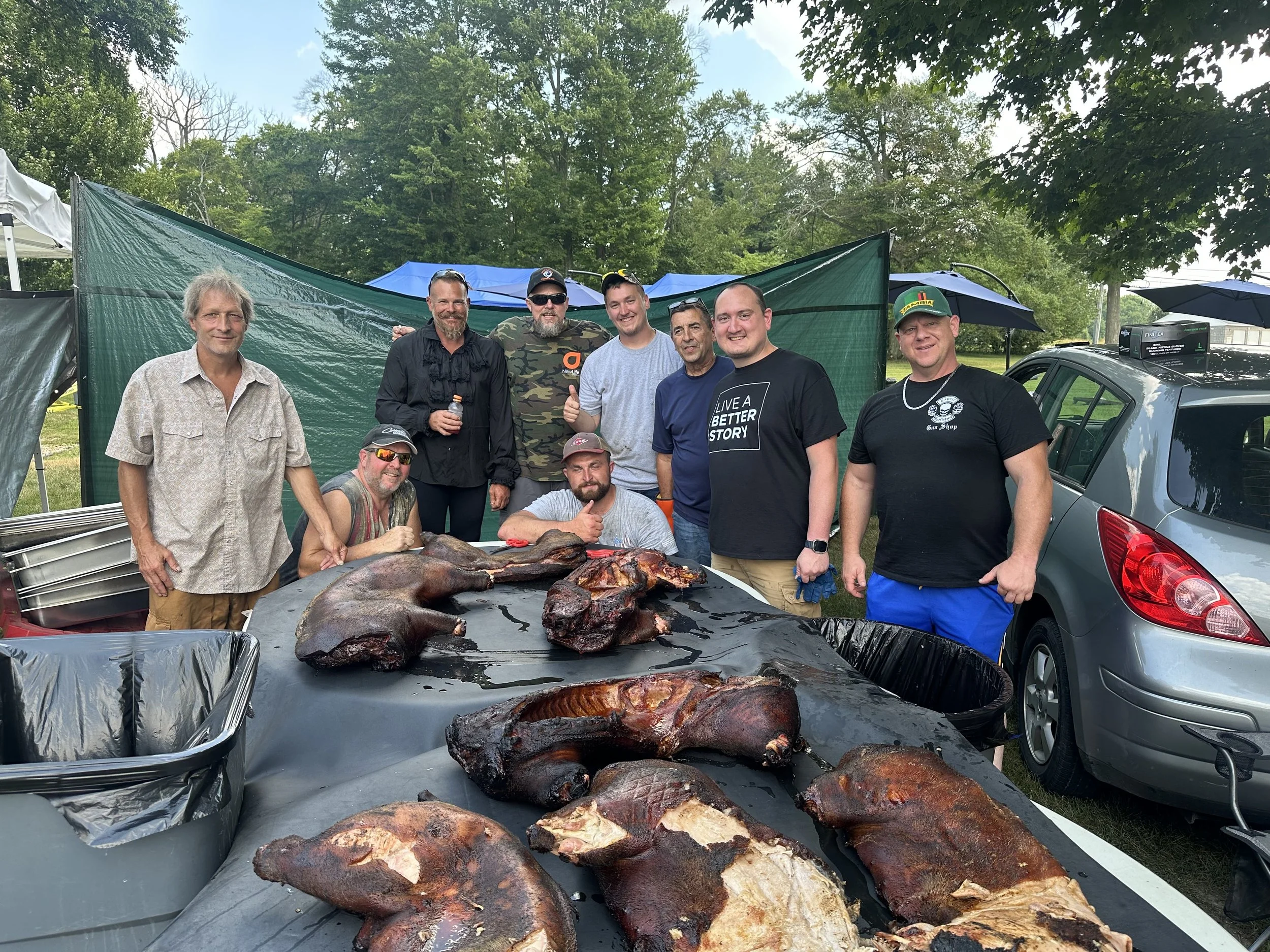 Group of men smiling and posing outdoors around a table with cooked fish, at a gathering with tents, trees, and parked cars in the background.