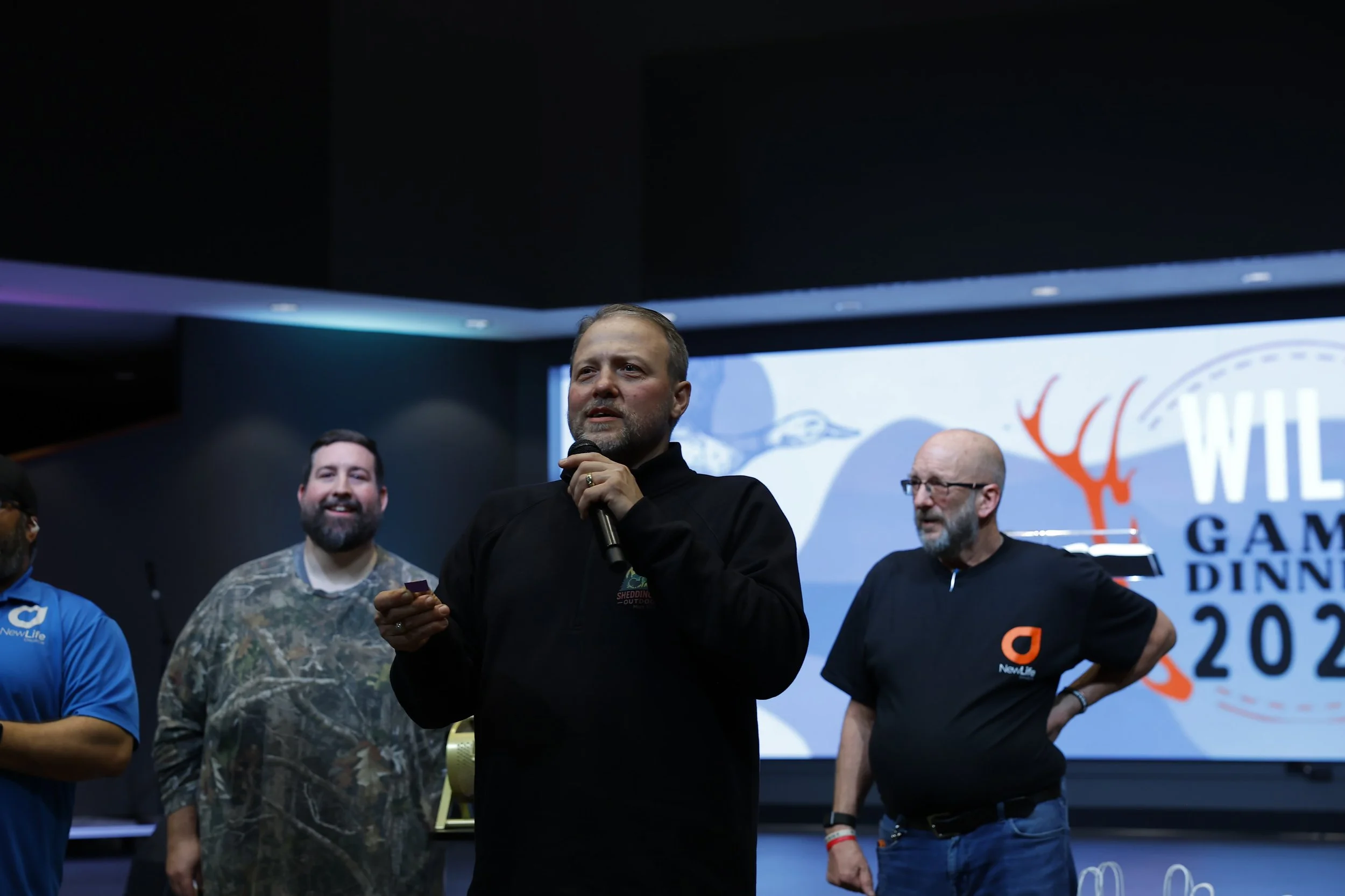A group of men standing on a stage at a conference, with a man in the center holding a microphone, during a presentation or panel discussion.