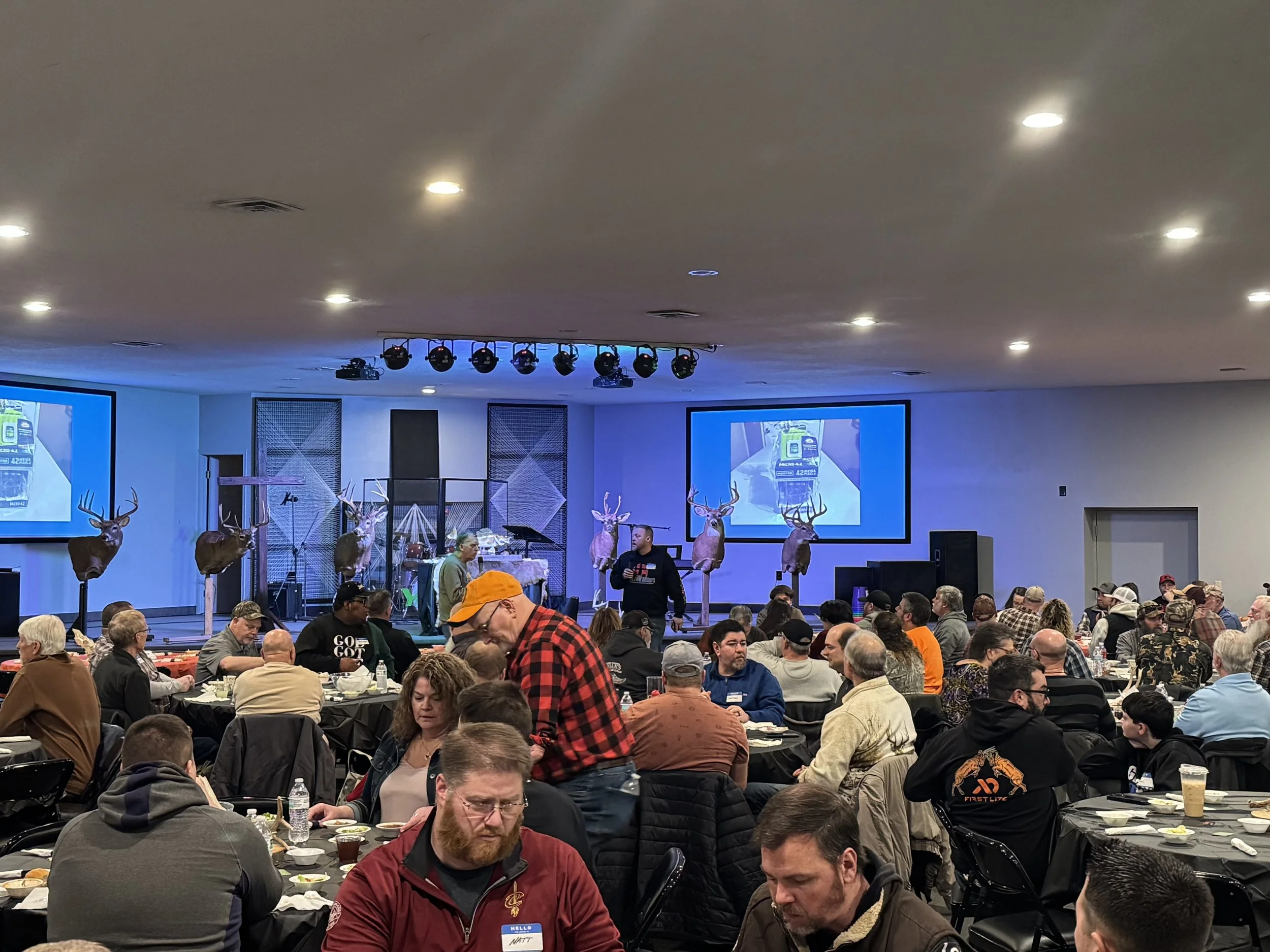 Indoor event with many people seated at tables, some with food and drinks, and decorative deer heads mounted on stands in the background. Large screens display a presentation, and there is stage lighting overhead.