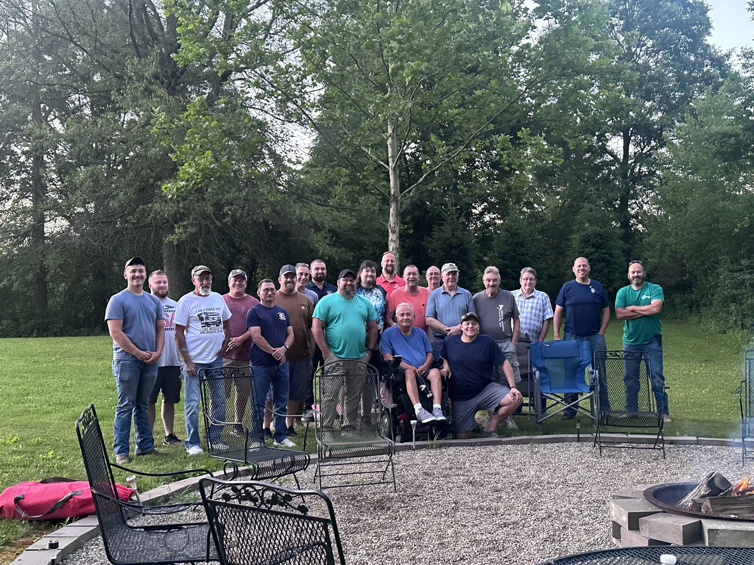 Group of people gathered outdoors in a park for a photo, with trees and a fire pit in the background.