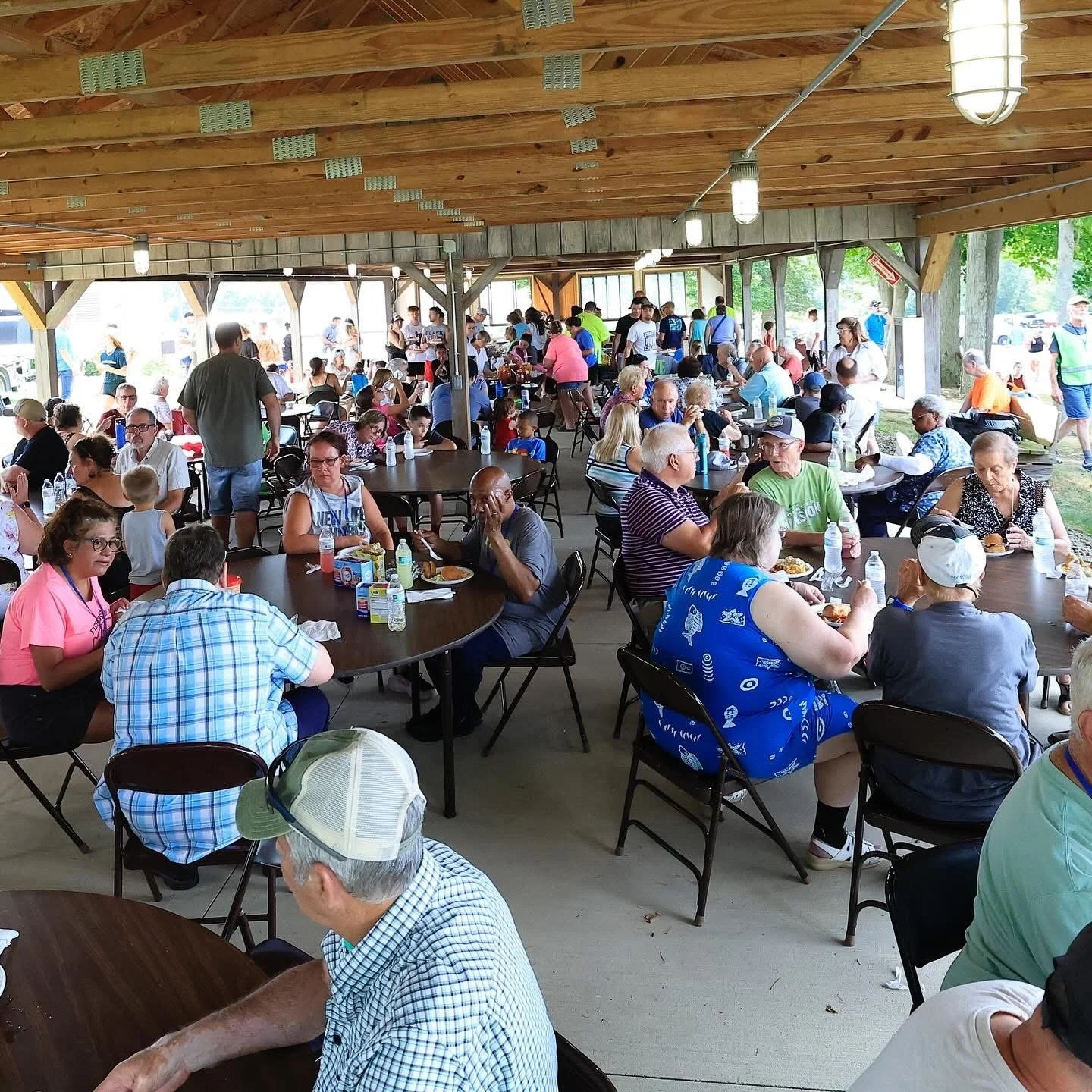 A large gathering of people sitting at tables under a pavilion at an outdoor event, eating and socializing, with more people standing and walking around in the background.