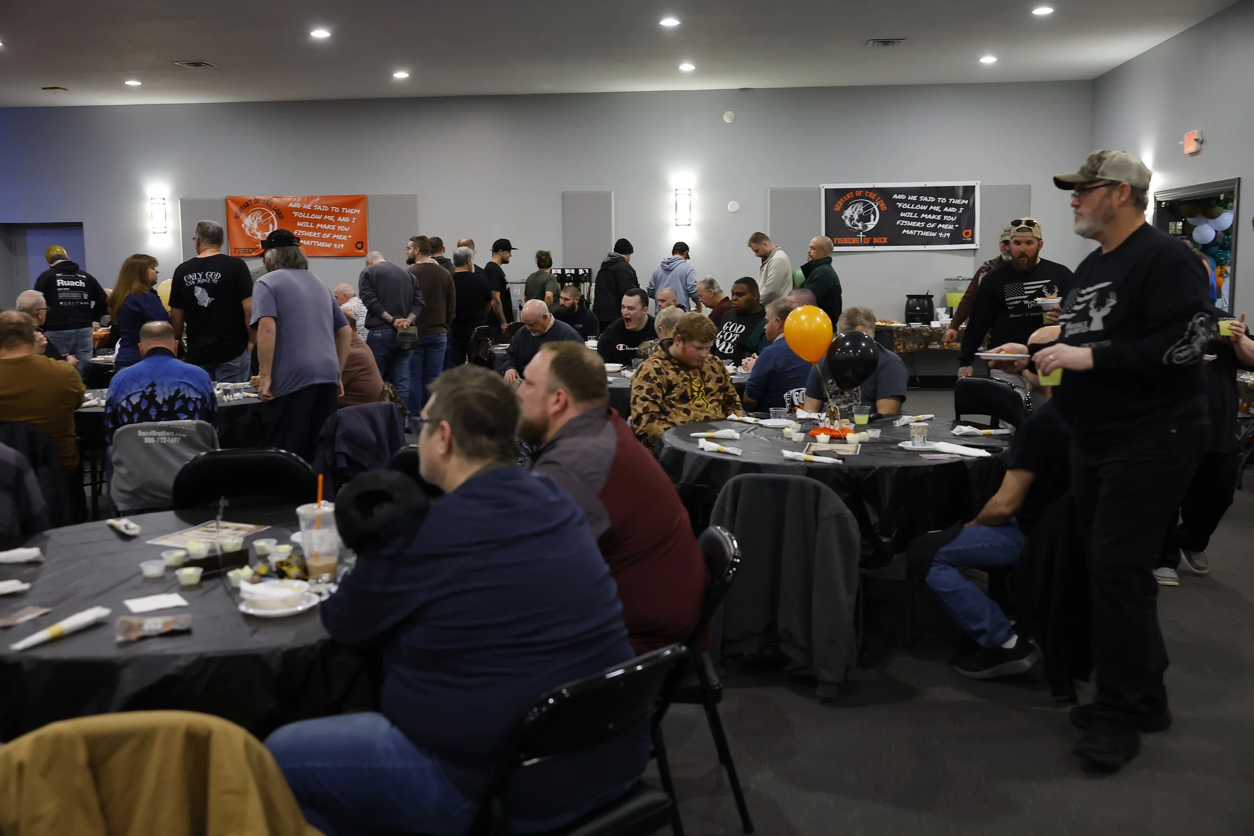 People gathered at a banquet or dinner event in a large hall, with tables set for a meal, decorated with balloons, and banners on the wall that display a religious or community message.