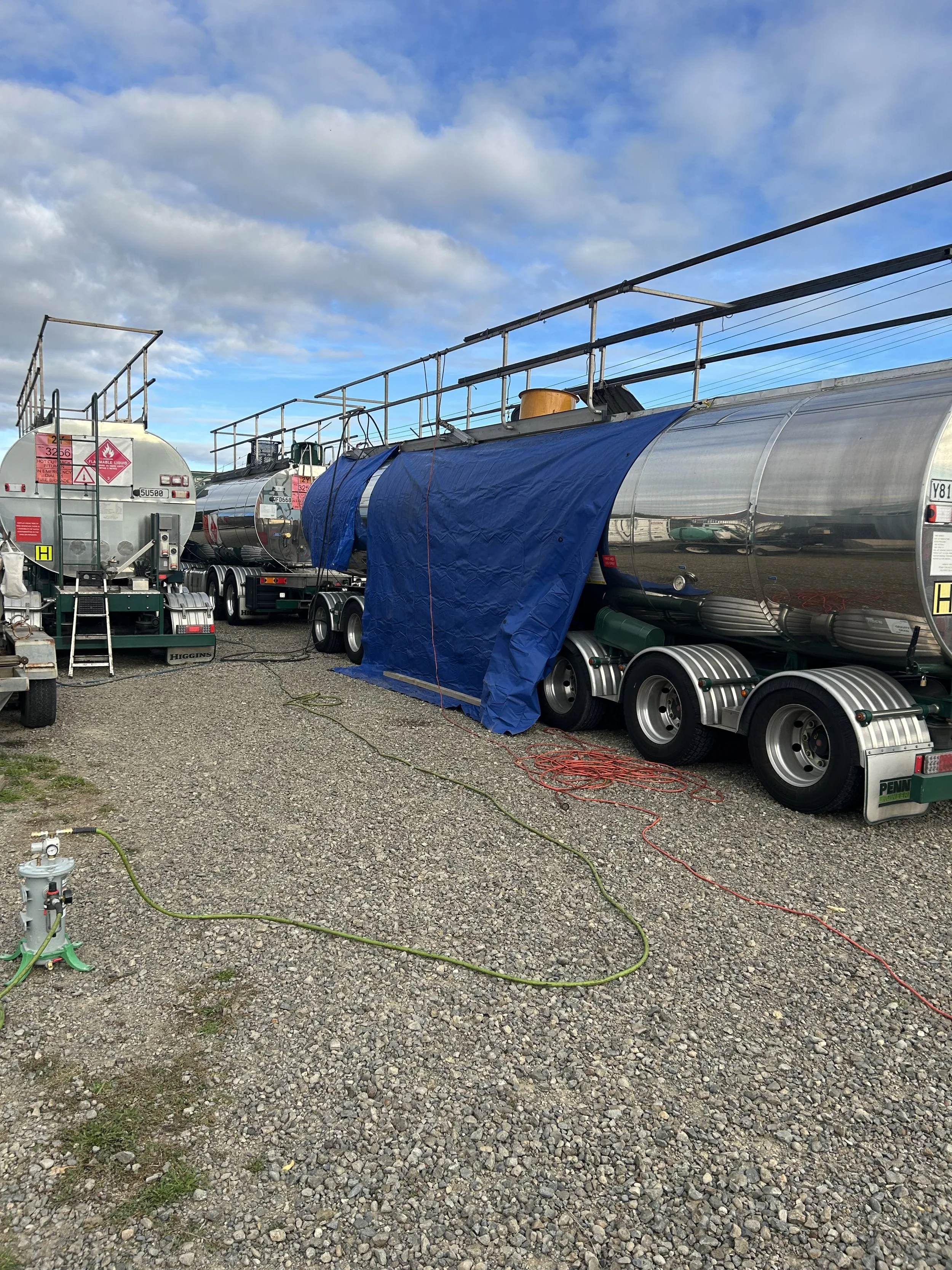 Several large bitumen tank trucks parked on a gravel lot under a partly cloudy sky, some covered with blue tarps, with hoses and equipment on the ground.