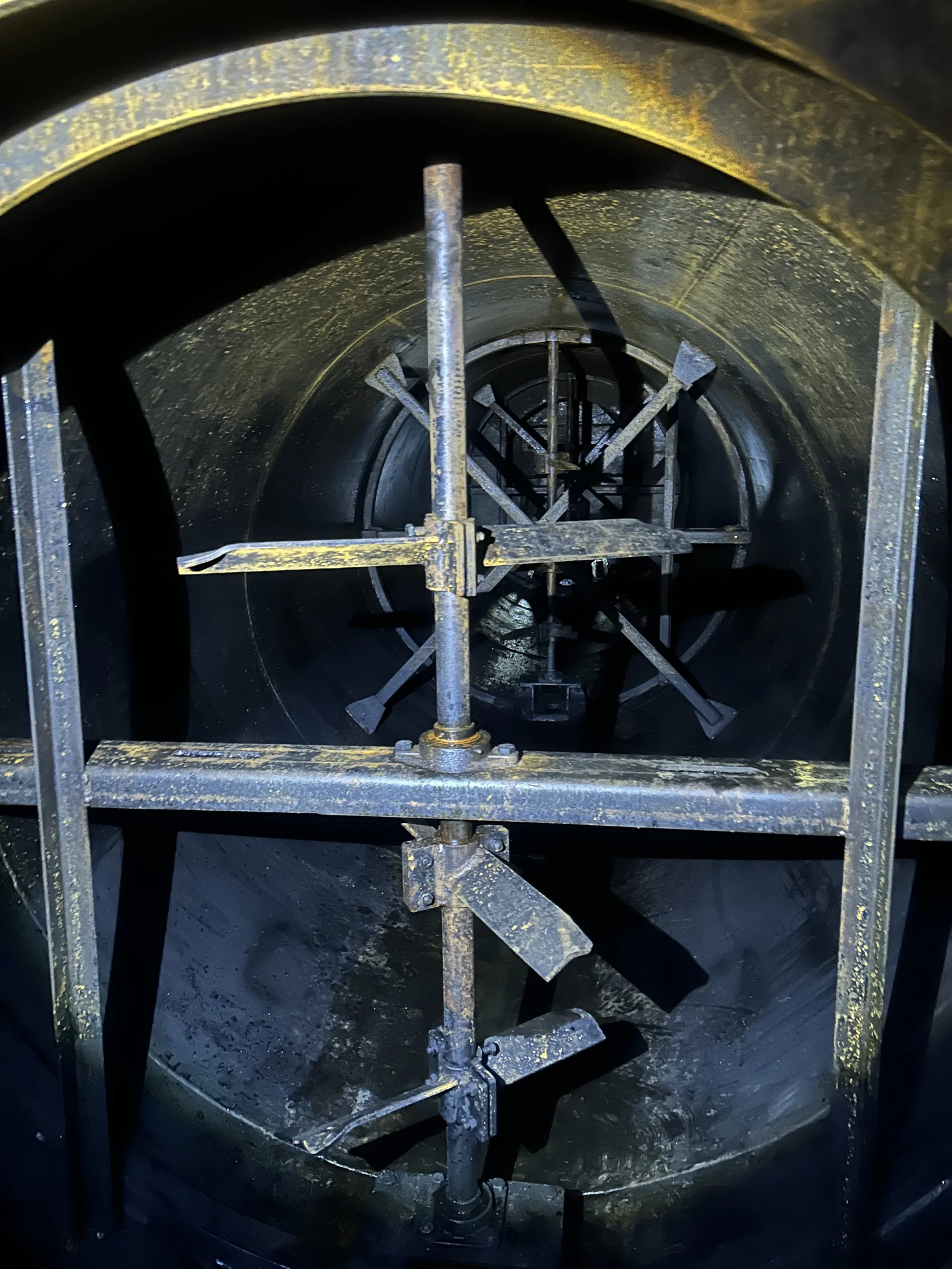 Inside a large industrial tank or pipe with metal scaffolding and support structures, showing a dark, cylindrical interior surface with some corrosion and grime.