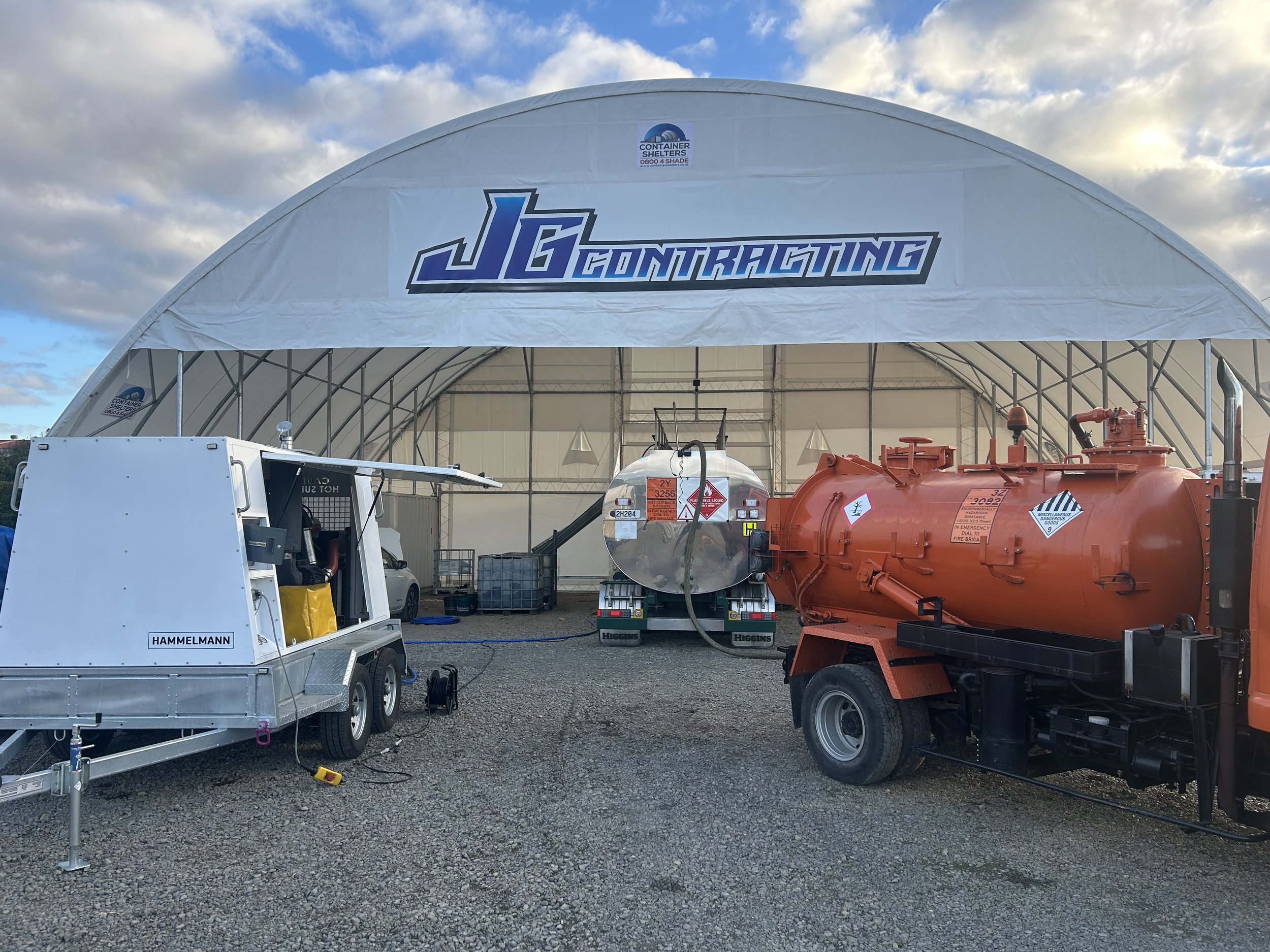Large white tent with 'JG Contracting' logo printed on it, surrounded by various equipment including a ultra high pressure water blaster, industrial vacuum truck, and bitumen tank.