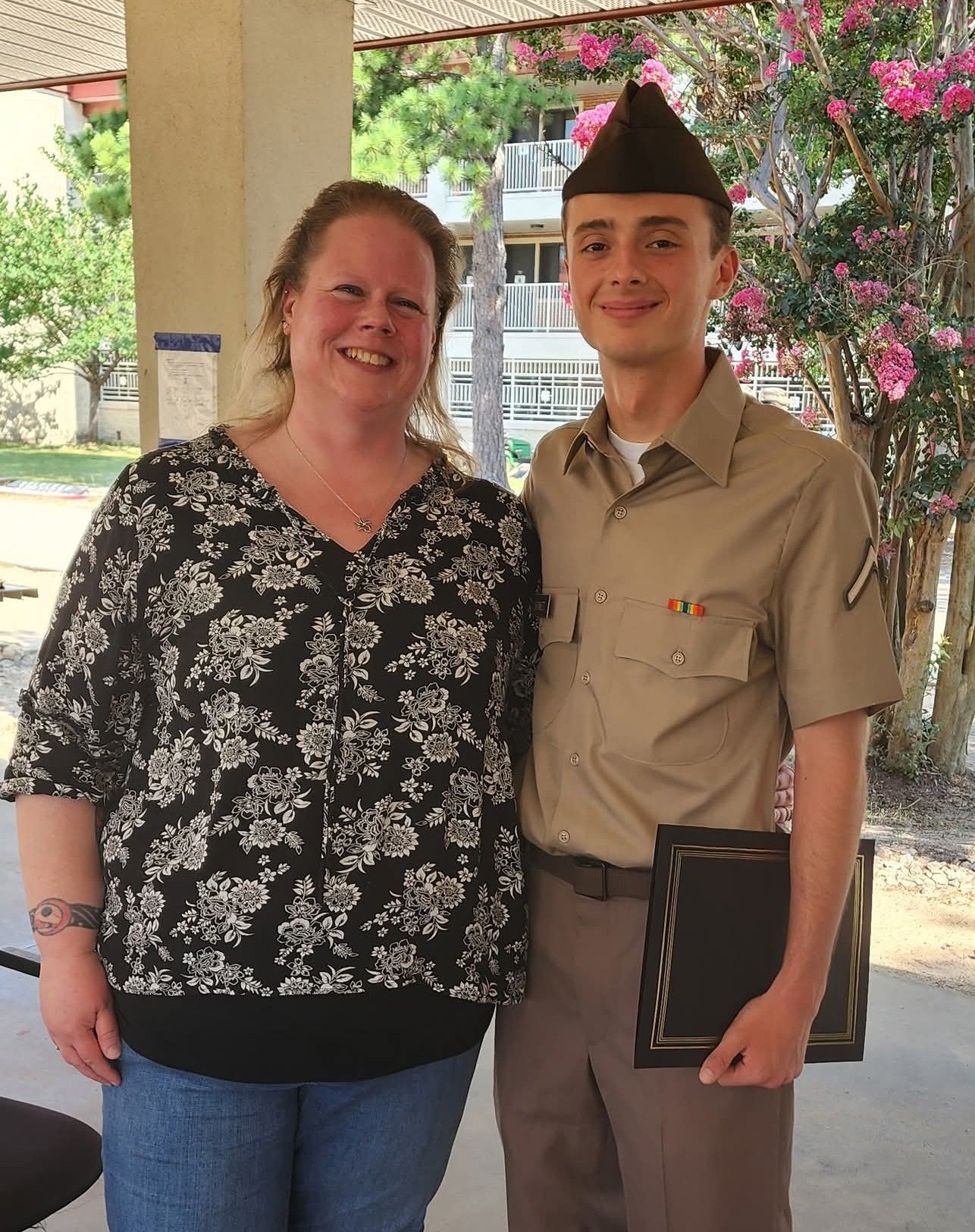A woman and a young man in a military uniform are smiling and standing together outside under a porch. The woman has long hair and is wearing a black and white floral blouse, and the young man is wearing a khaki uniform with a ribbon bar on his chest, and a brown military hat. There are pink flowering bushes and trees in the background.