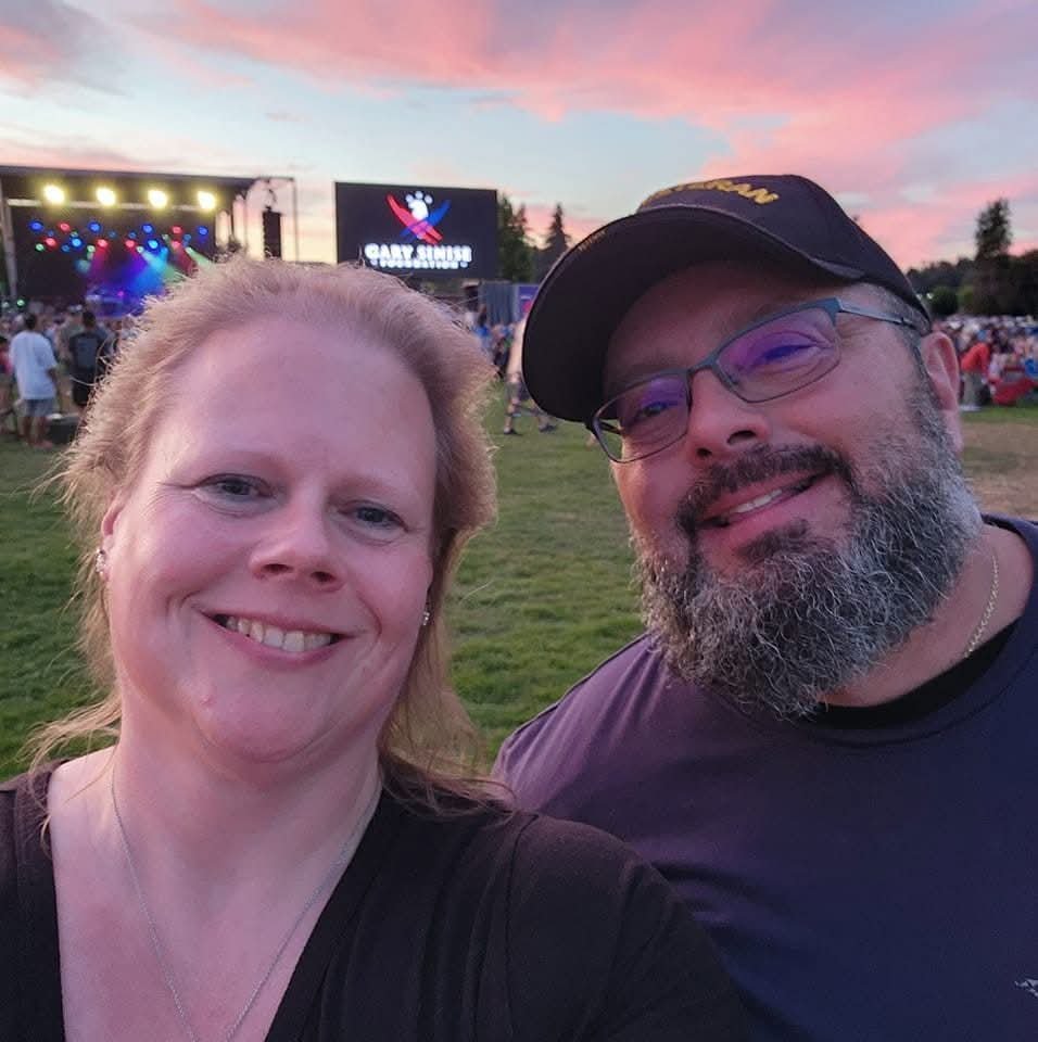 A smiling woman and man taking a selfie outdoors during a concert at sunset, with the stage and audience in the background.