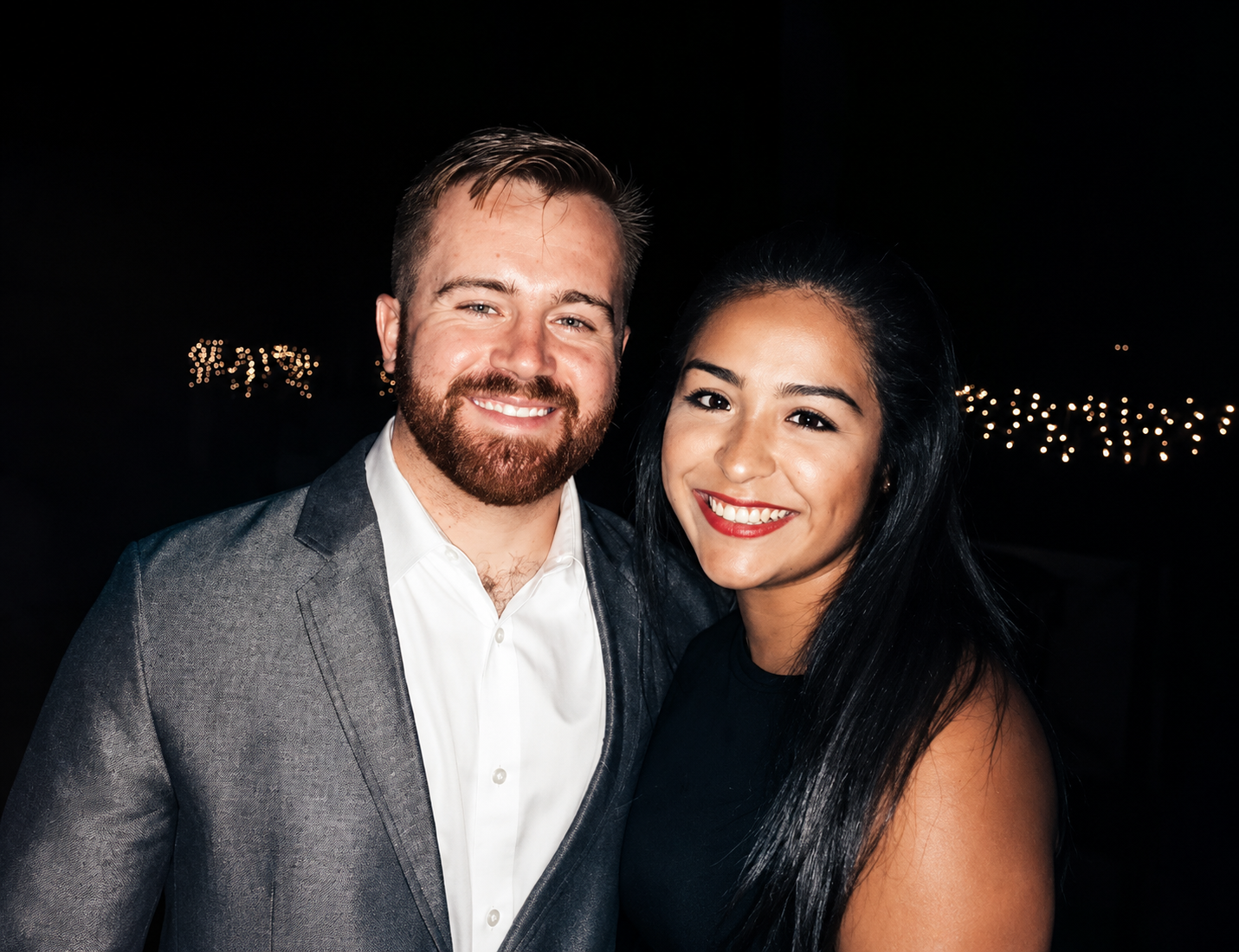 A smiling man with a beard in a gray suit and white shirt standing next to a smiling woman with long black hair and red lipstick in a black dress, at night with string lights in the background.