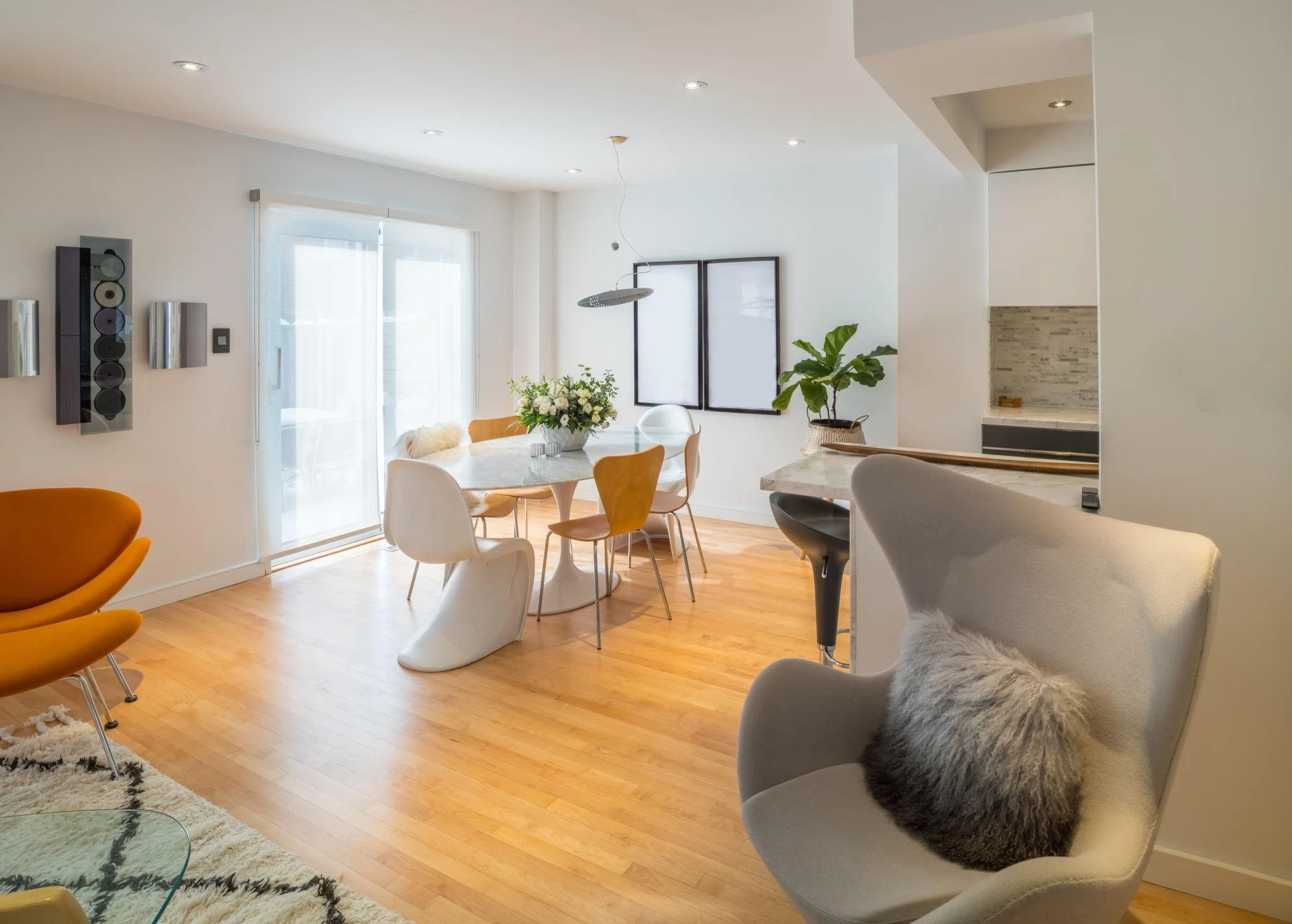 Modern living room with white walls, wooden floor, and large glass doors allowing sunlight. Includes a dining table with flowers, stylish chairs, a cozy hanging chair with furry blanket, potted plant, and contemporary wall decor.