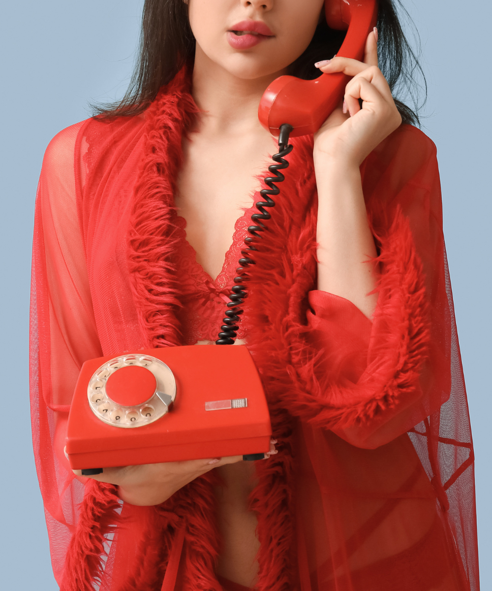 A woman wearing a sheer red robe holding a vintage red rotary telephone to her ear.