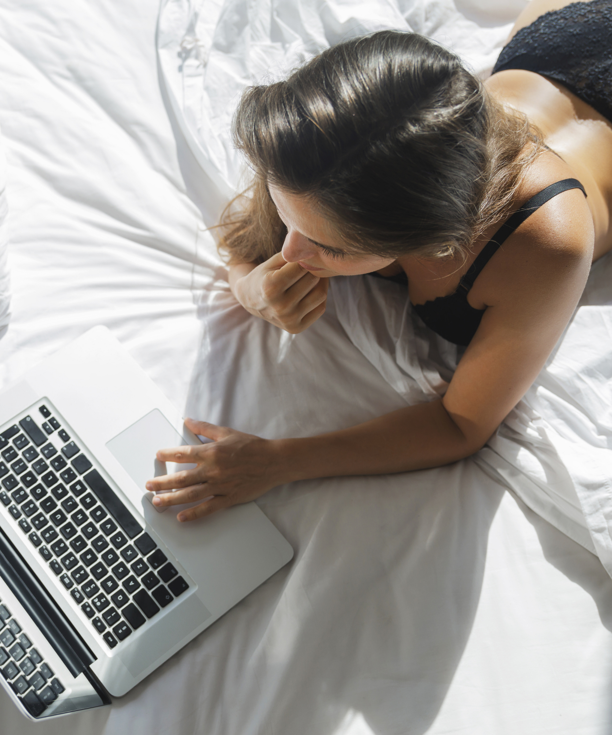 A woman with brown hair, wearing a black tank top, lies on a white bed using a silver laptop. She is resting her head on her hand and appears to be engaged with her device.