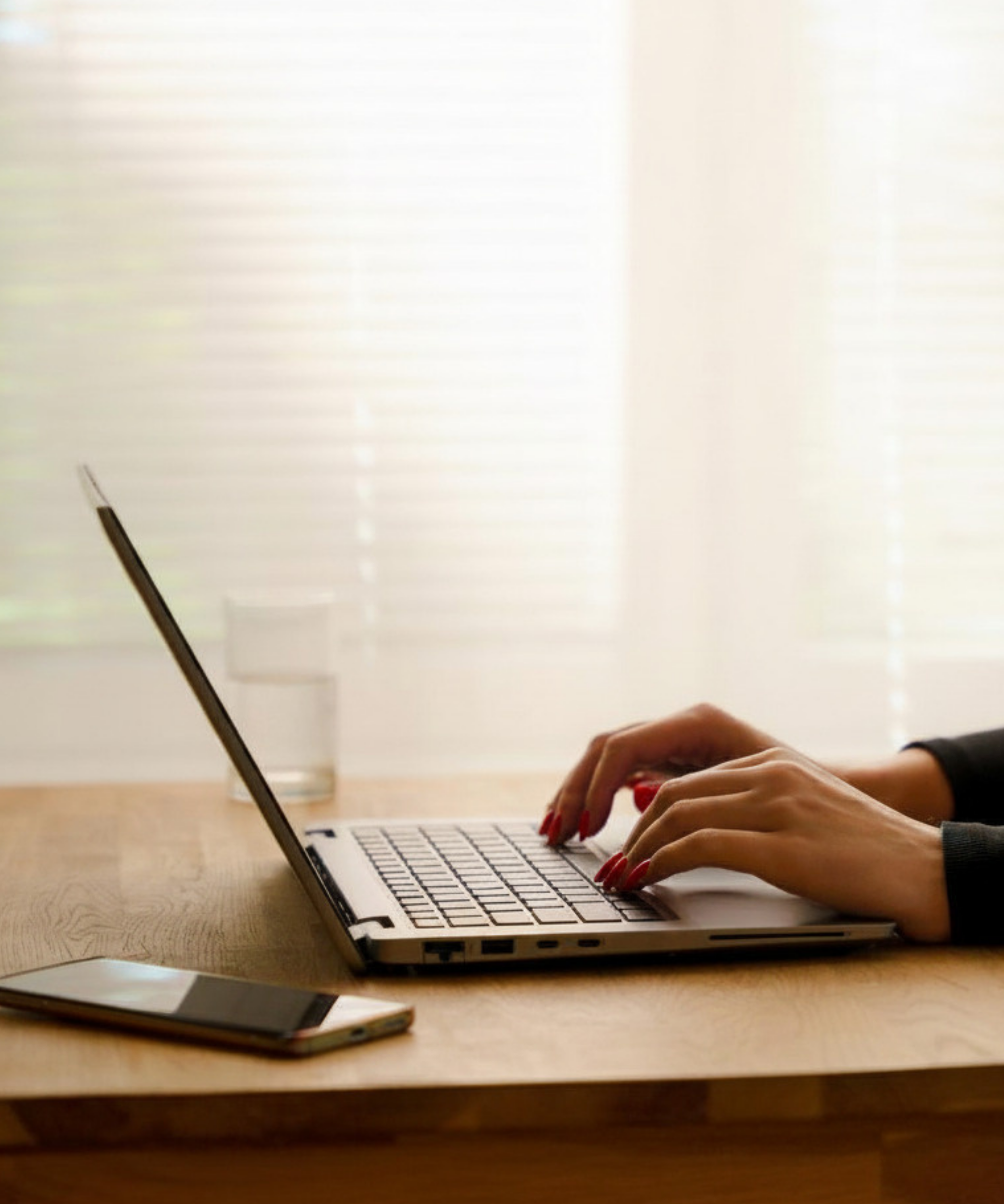 Person typing on a laptop computer with a mobile phone and glass of water on a wooden table.