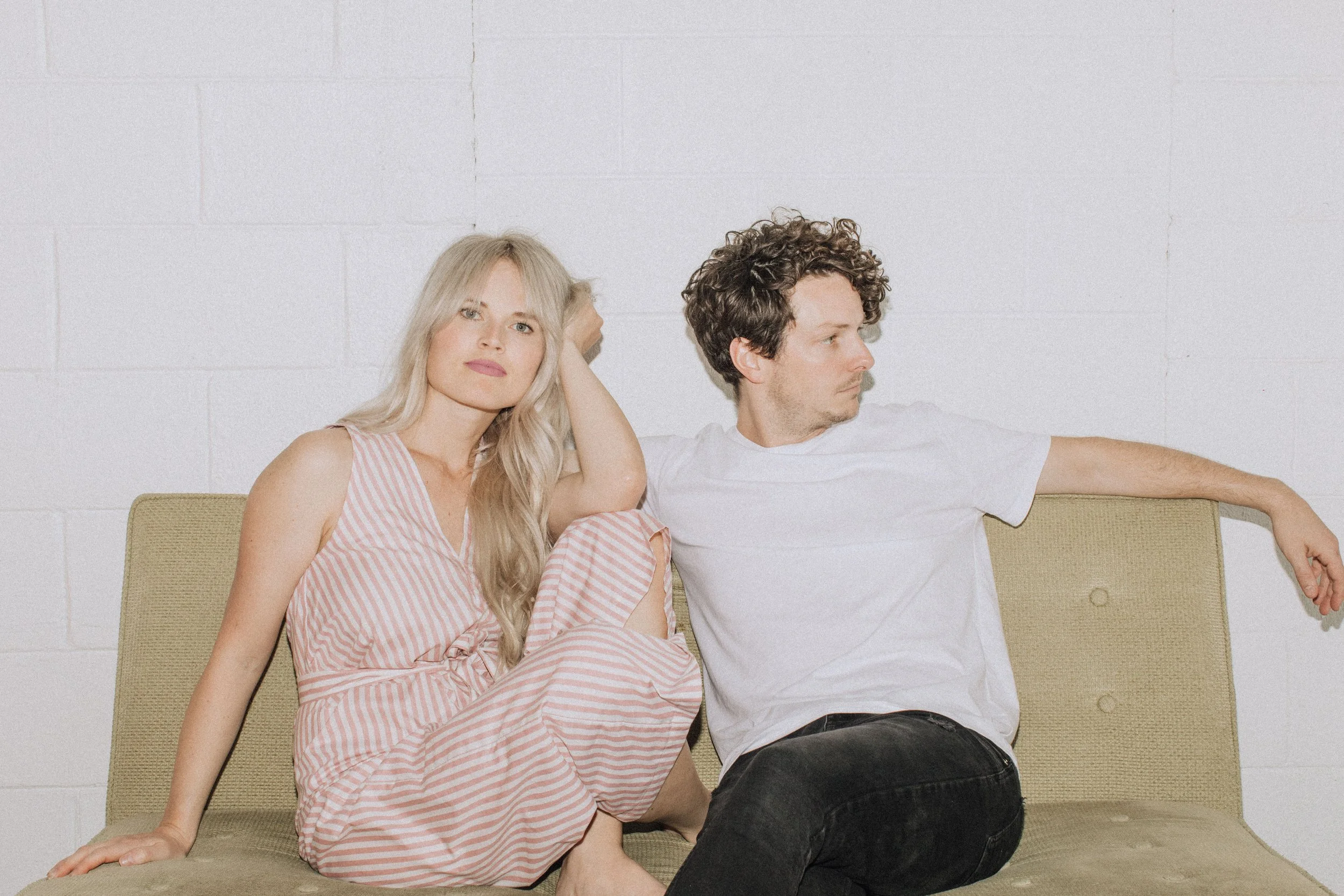 A young woman with long blonde hair wearing a pink and white striped dress sitting on a beige couch next to a young man with curly brown hair wearing a white t-shirt and black pants, against a white brick wall background.