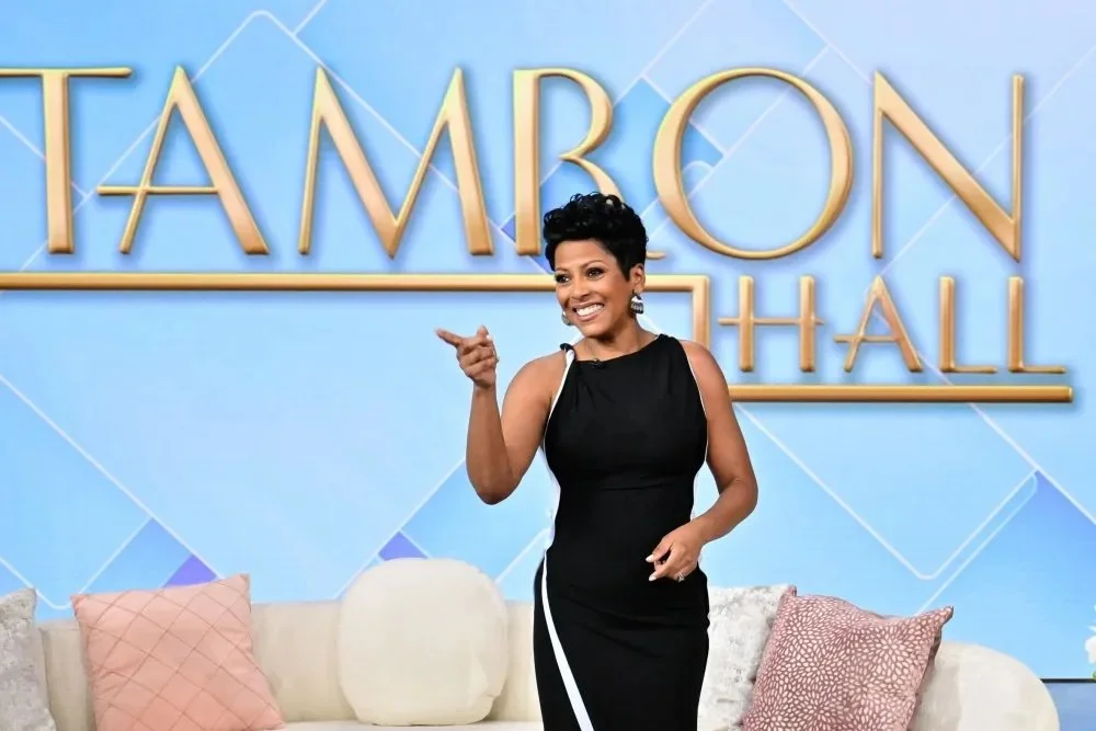 A woman with short black hair wearing a black dress with white accents, smiling and pointing, standing in front of a blue backdrop with large gold letters spelling "TAMRON HALL". There are pink and white pillows on a beige couch nearby.