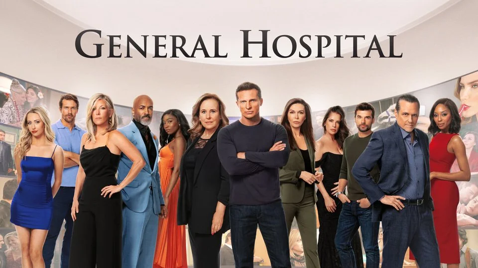 A group of eleven diverse people standing in front of a wall with 'General Hospital' written on it, in a hospital reception area, posing for a photo.