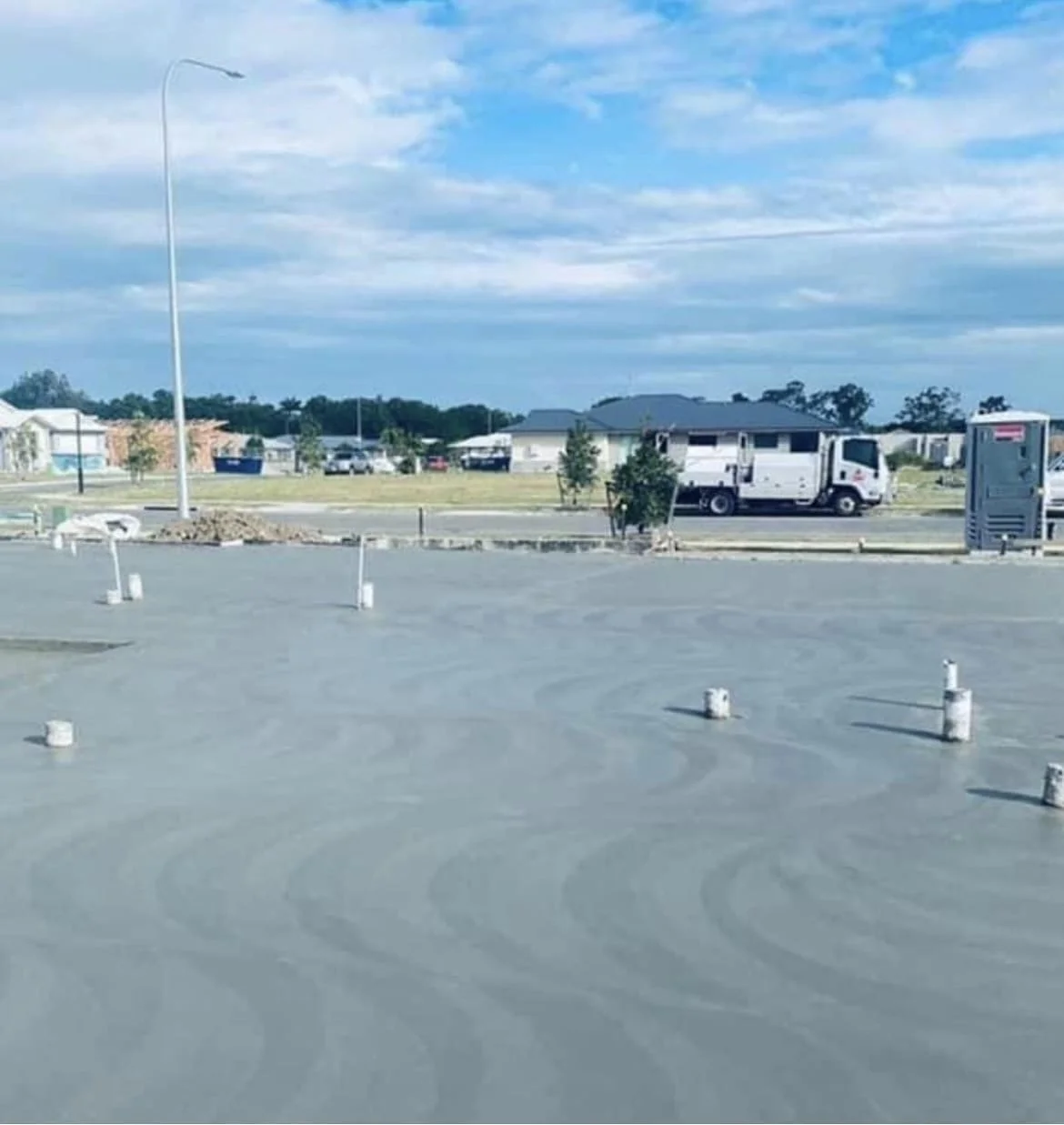 View of a construction site with a smooth, level surface, small white posts, and a pickup truck in the background under a partly cloudy sky.