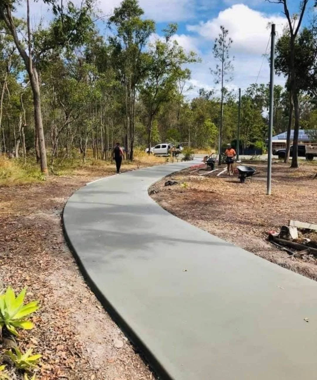 Newly poured concrete sidewalk under construction on a curved road section with construction equipment and dirt piles in the background.