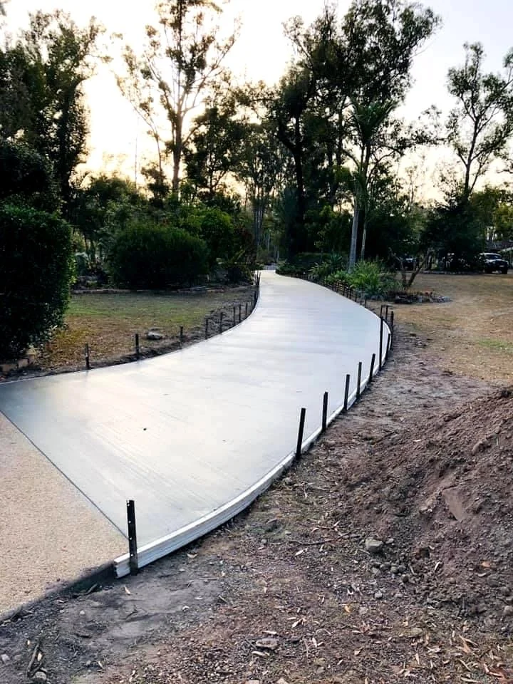 Newly constructed concrete pathway with black metal stakes along its edges, curving through a park or garden area with trees and bushes.