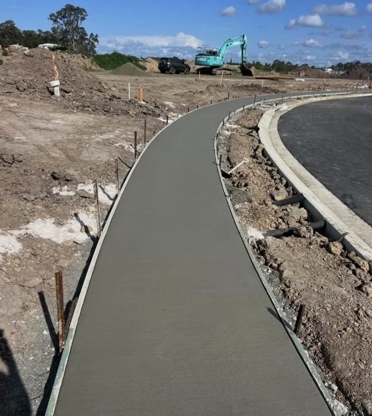 Newly poured concrete sidewalk under construction on a curved road section with construction equipment and dirt piles in the background.