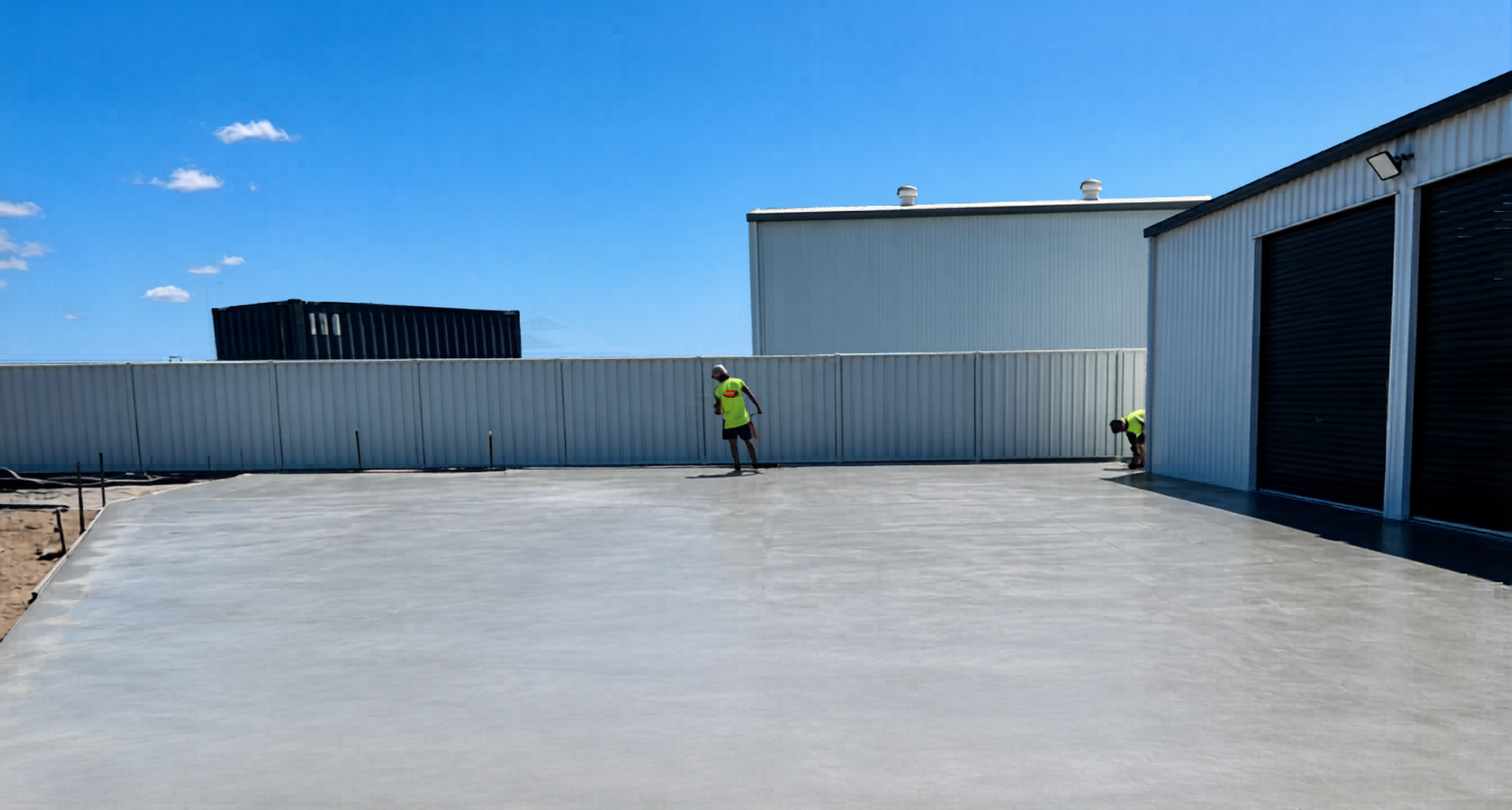 Construction workers in high-visibility yellow shirts working on a freshly poured concrete surface outside a building with metal walls and rolling doors under a clear blue sky.