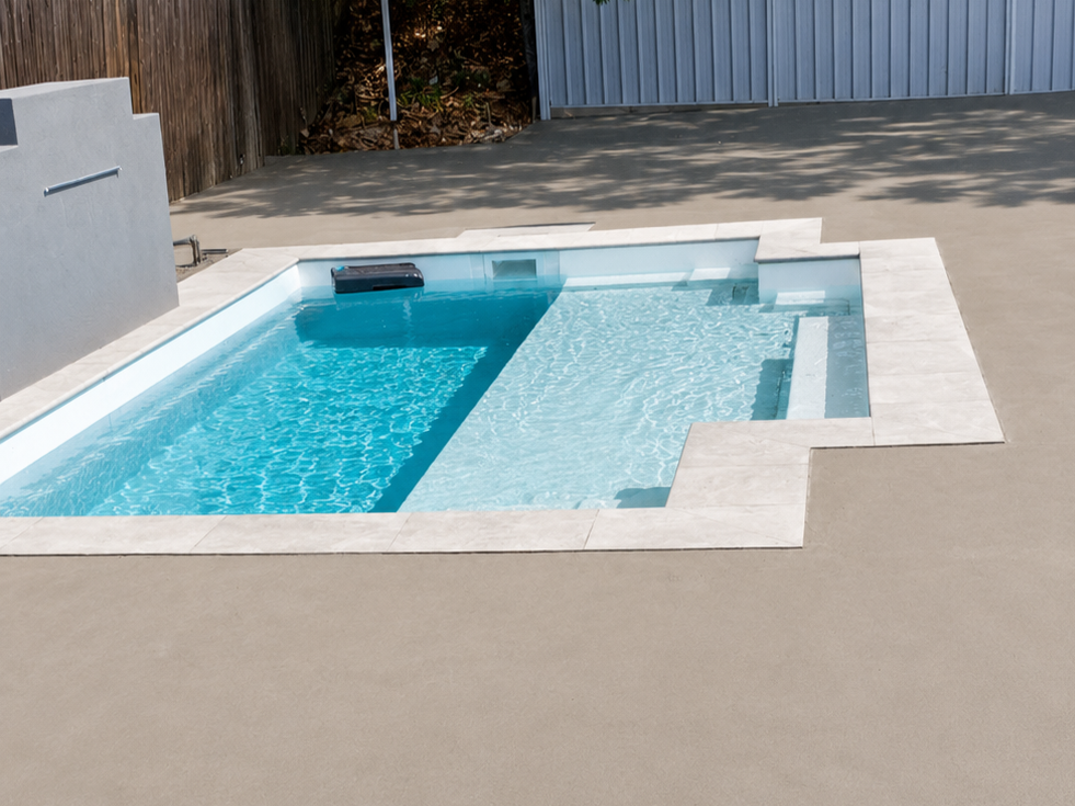 A small in-ground swimming pool with a spa section, surrounded by a concrete deck, with a wooden fence and a metal fence in the background.