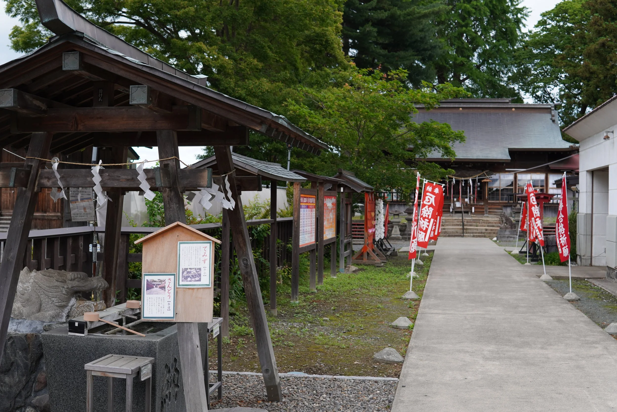 神社の入り口に木製の鳥居と旗が立ち並び、木造の社殿と緑の木々が背景に見える静かな境内の風景.