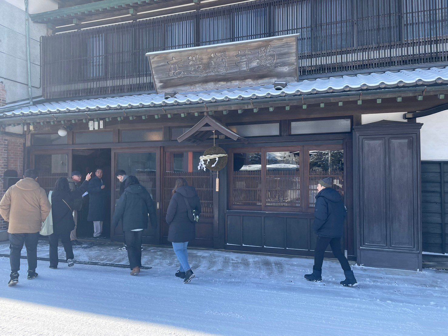 雪のある路地に古い木造の店の前に人々が並んでいる。店の看板には日本語で「酒井醸造店」と書かれている。